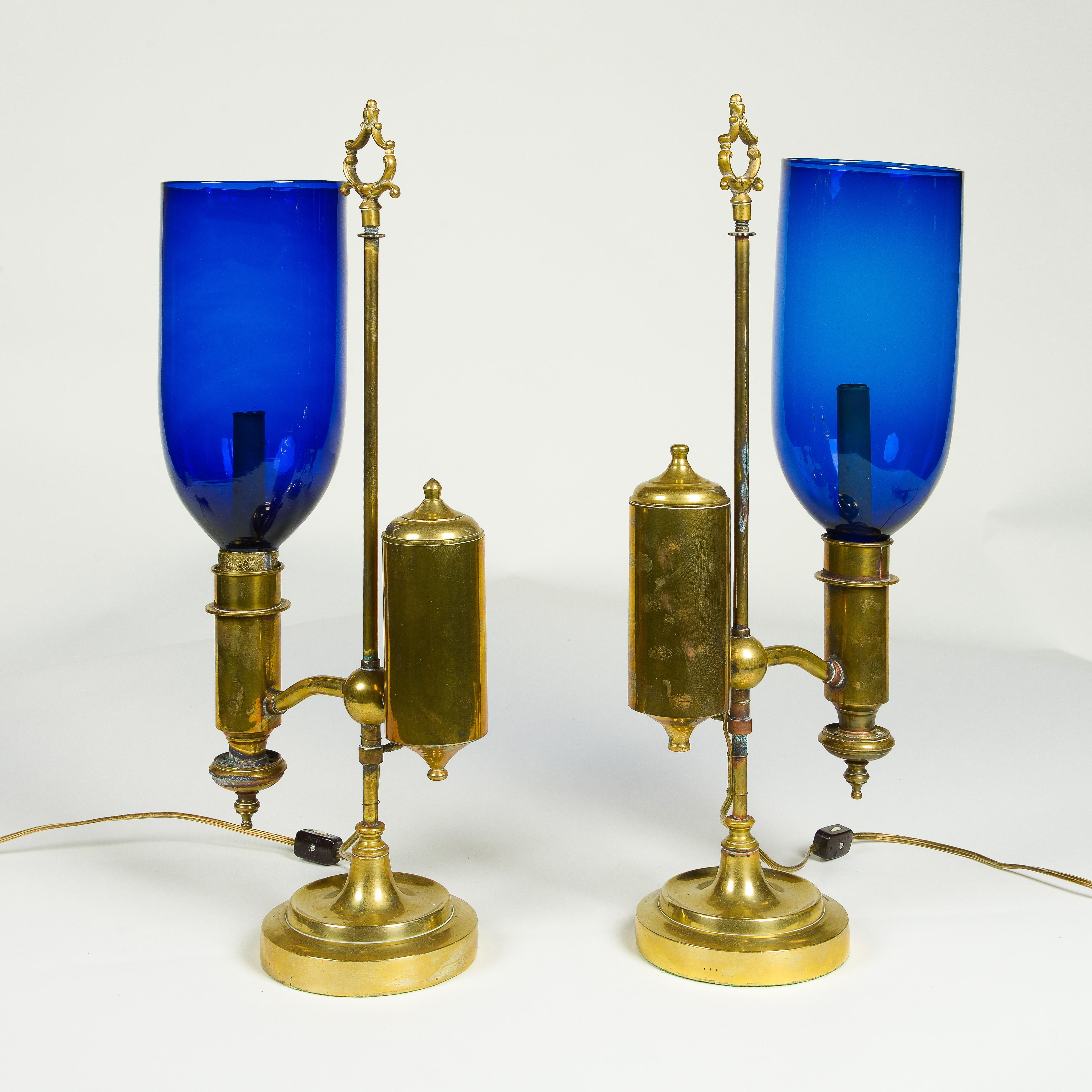Each oil lamp later covered to electric; fitted with an adjustable arm issuing one light with later, removable blue hurricane shade; surmounted by a removable finial in the form of an open scrollwork clasp.