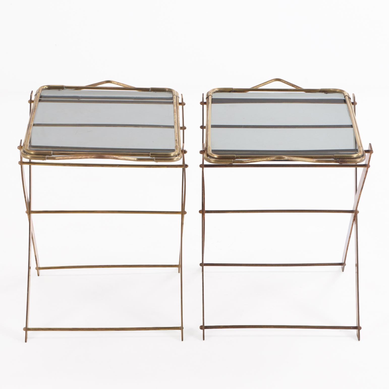 Questa elegante coppia è caratterizzata da vassoi rimovibili con superfici in vetro fumé, sostenuti da una base pieghevole in ottone per un uso versatile. Il vetro colorato aggiunge un tocco sottile e sofisticato, mentre la cornice in ottone dona