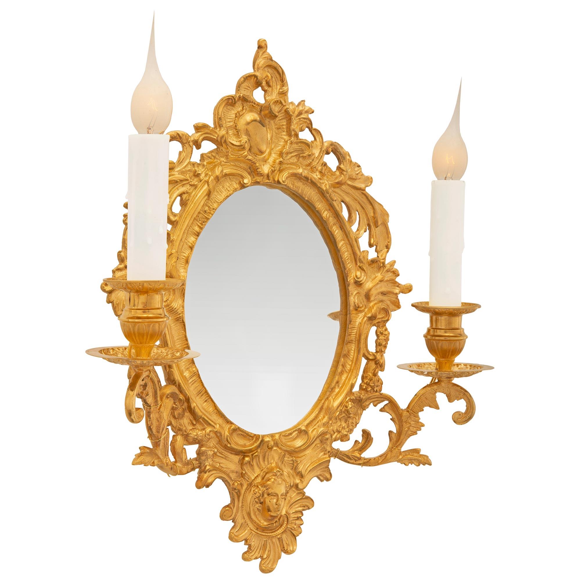 st. Ormolu mirrored sconces. The pair of two arm sconces have pierced oval backplates surrounding the central mirror plate. The frame has a bottom central reserve of a female face while the sides of the frame display pierced foliate scrolls with