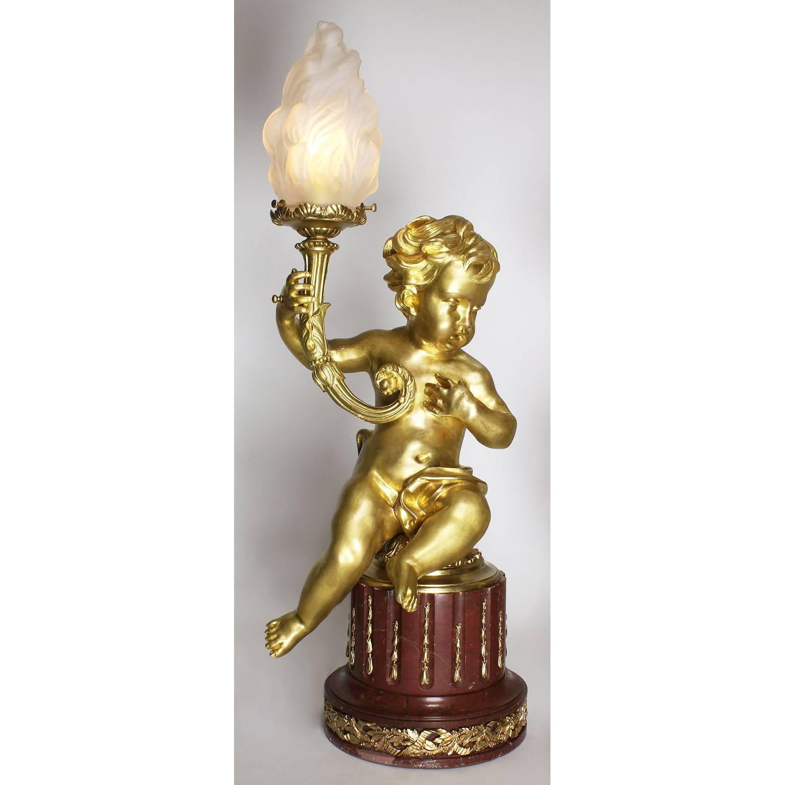 A fine pair of French 19th-20th century Louis XV style figural gilt bronze Putti torchere table lamps on a Rouge Royal base. Each figure representing a seated Putto (child) holding a scrolled gilt bronze flambeaux fitted with a frosted glass shade