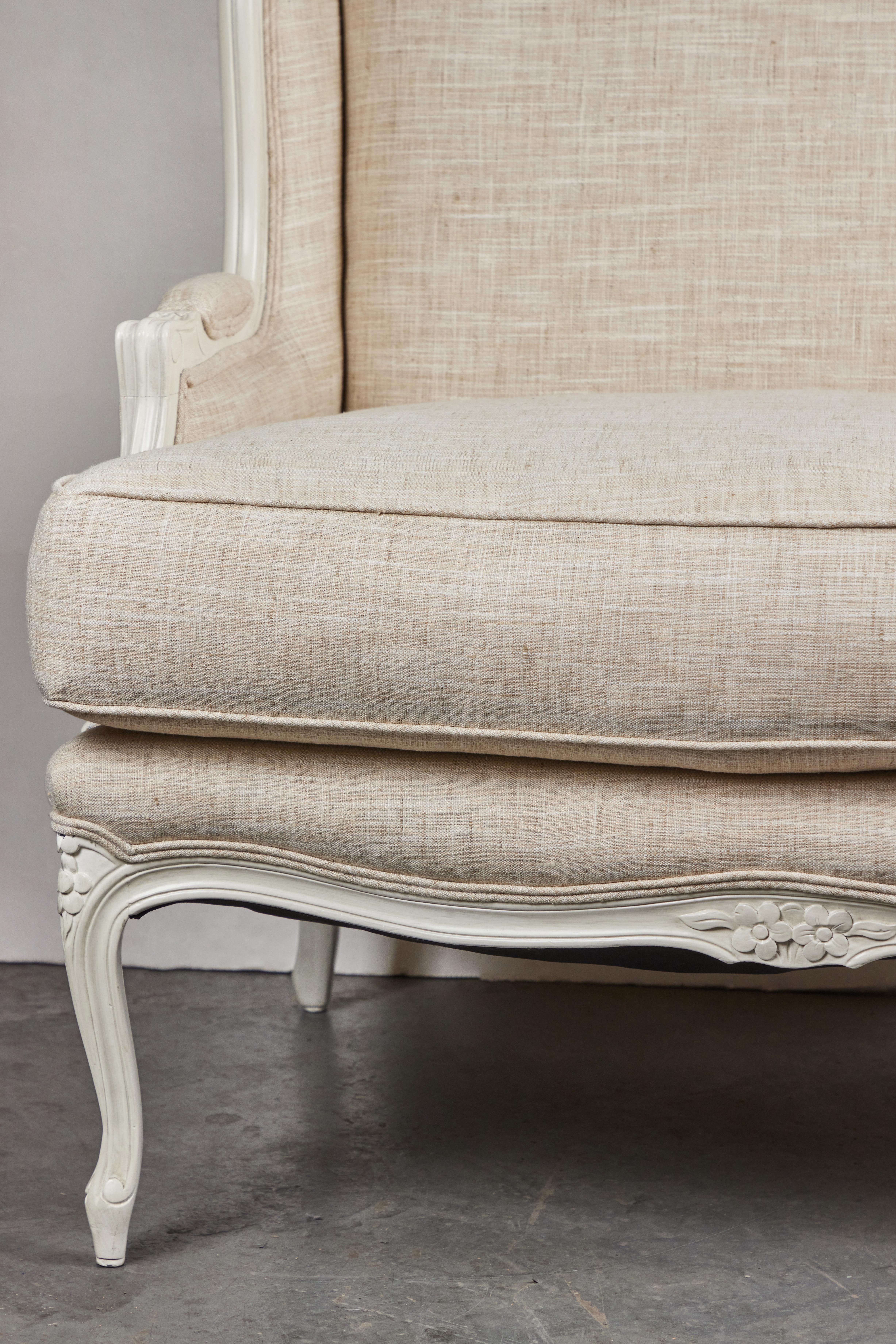 An elegant pair of French bergères in crème paint and new linen. Each with floral relief top rails, and resting on cabriole legs.