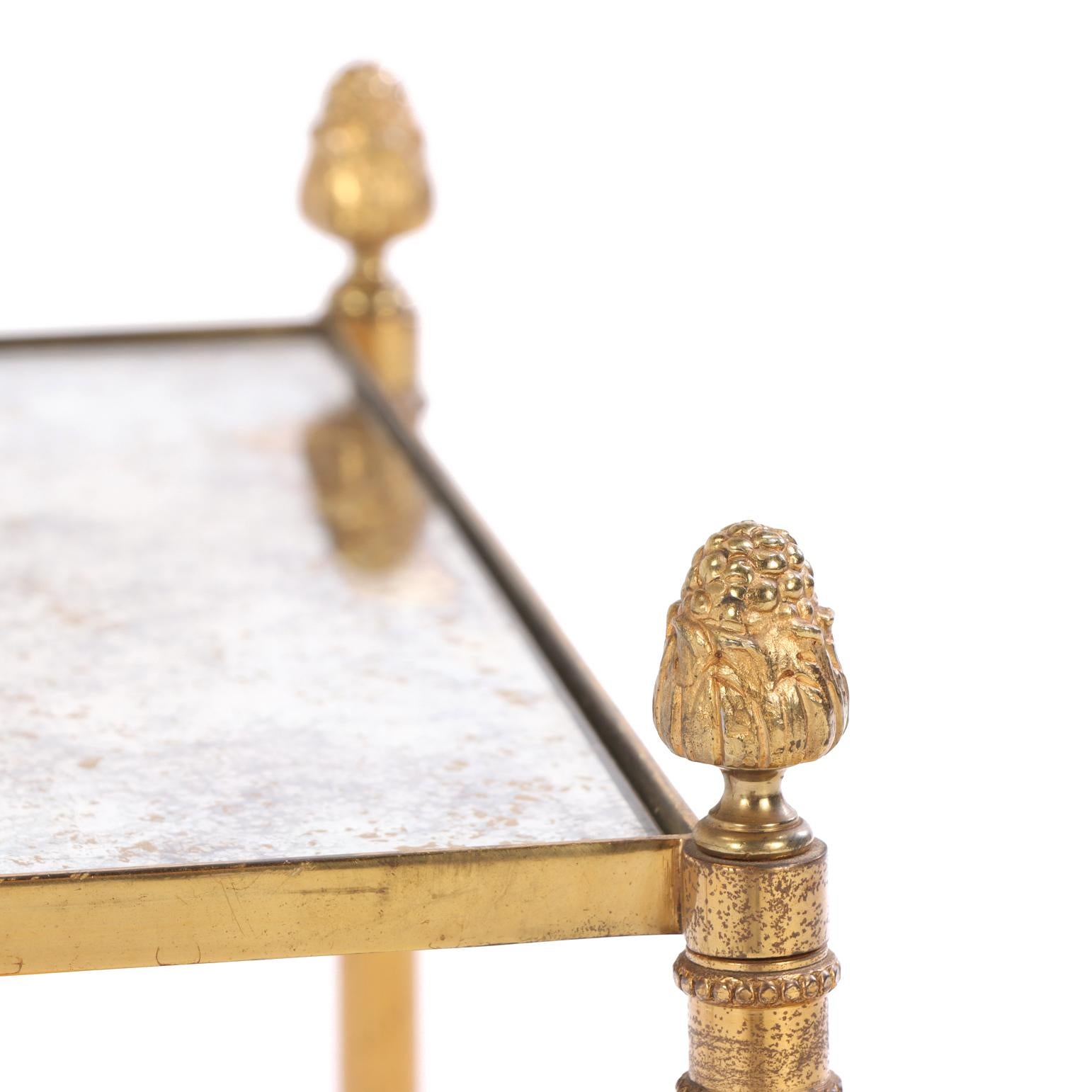 Each table features a square mottled mirrored top inset within a brass frame, supported at the corners by four uprights surmounted by decorative acorn finials. The bases are united by an X-form stretcher centered by a matching finial.

The design