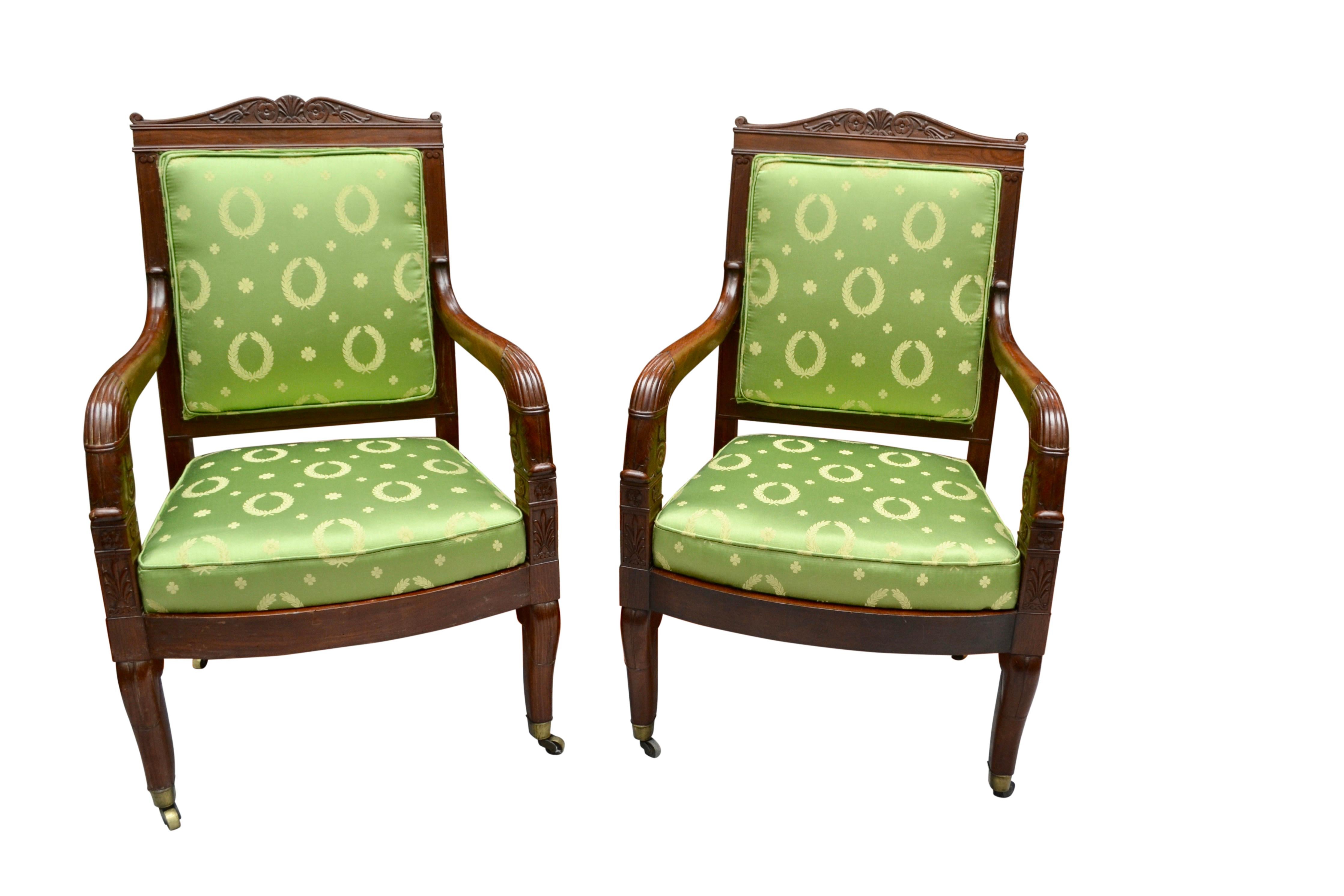 A classic pair of French Empire period open armchairs in well figured and carved mahogany; each with a square back topped by a bow shaped carved frieze featuring folliate rosettes and downscrolled arms. Where the arms join the chair rail there are