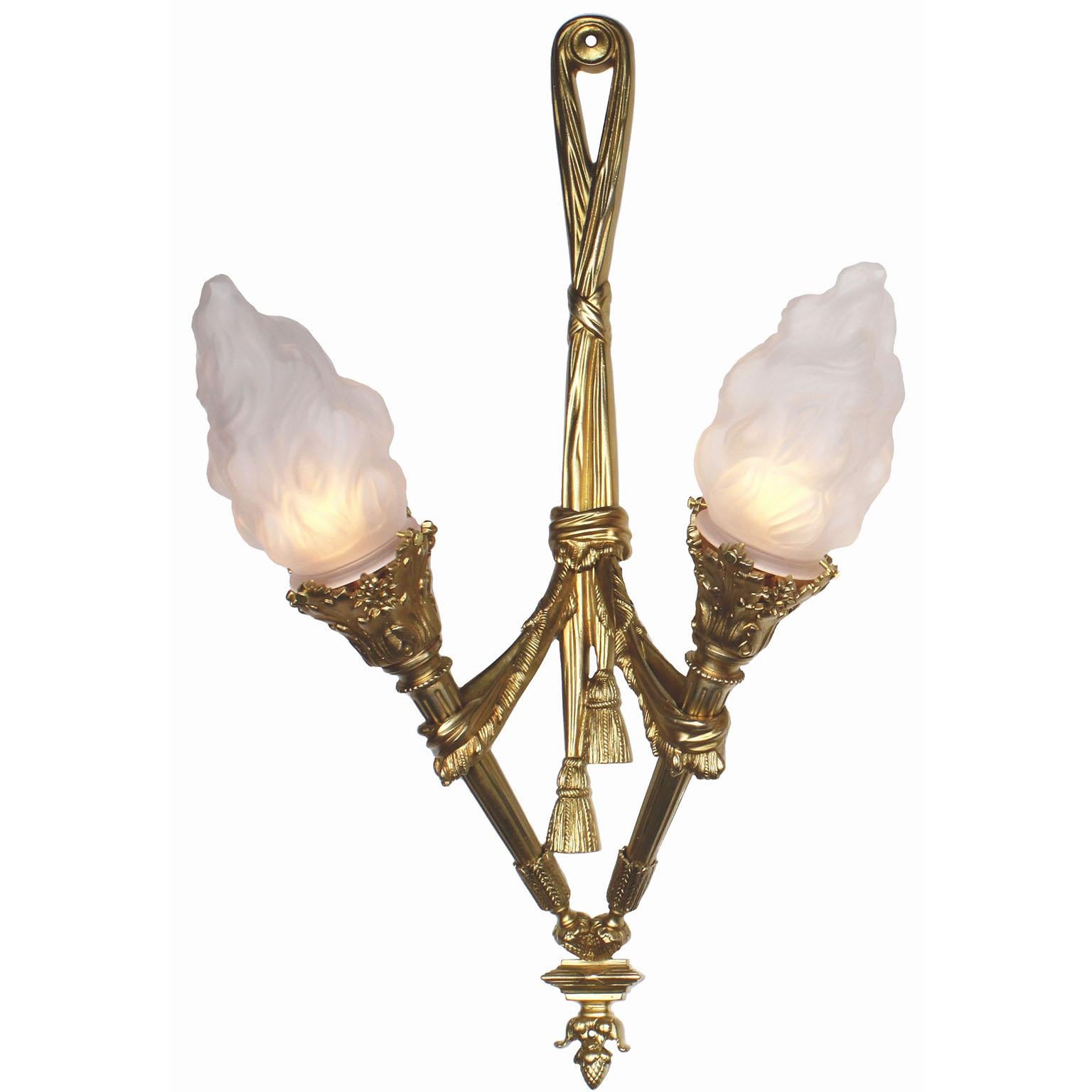 A pair of French empire style gilt-bronze two-light wall torchère sconces. Each elongated gilt-bronze applique with central twin-tassels supporting a pair of torches, each fitted with frosted glass flame shades and ending with acorn finials.