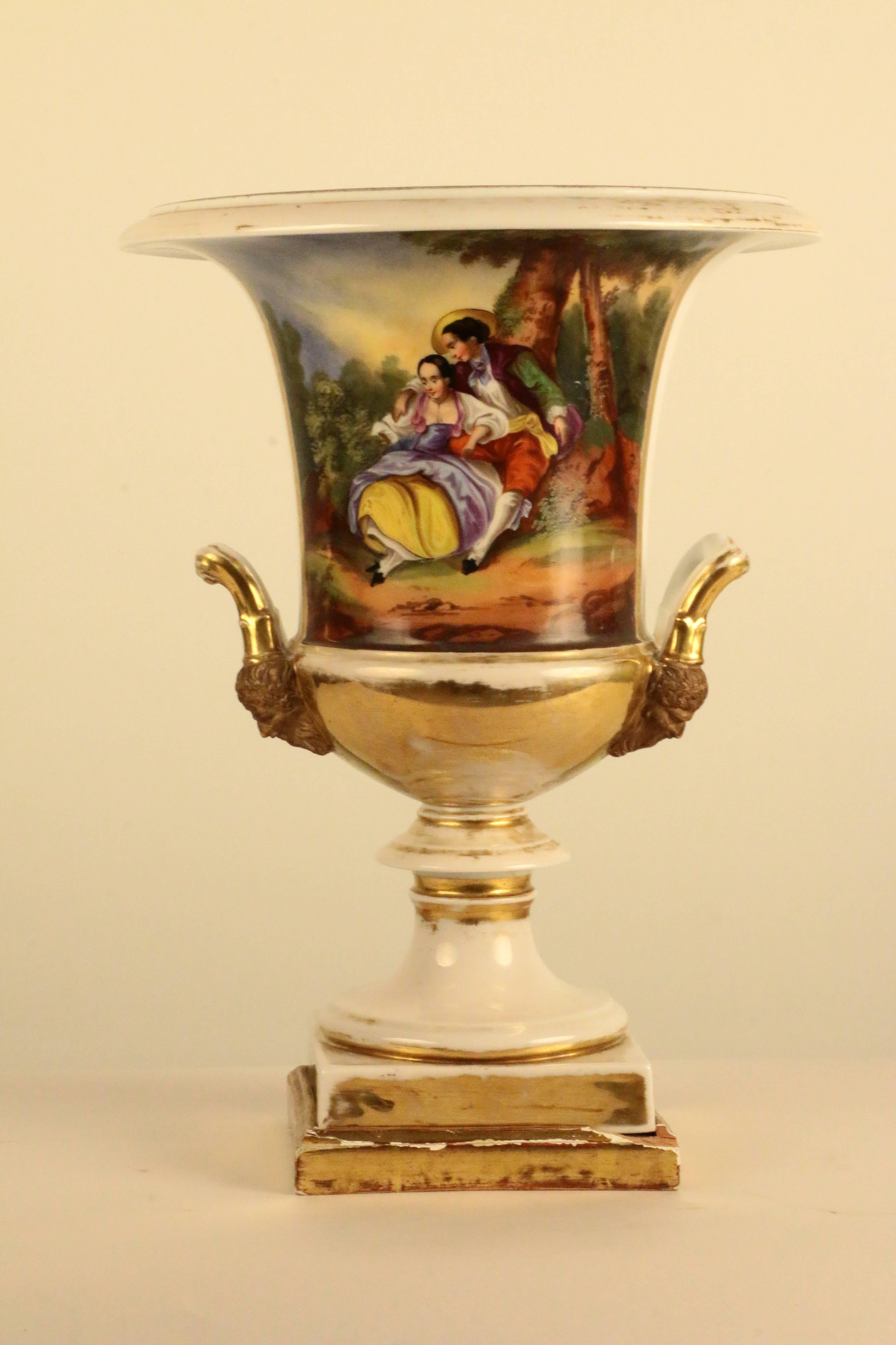Now fitted as lamps, these two handled Paris urns are painted with floral bouquets on one side and a courtship scene on the other. The gilding is rubbed but the painting is well executed and a charming subject. Each is lit by a single candleform