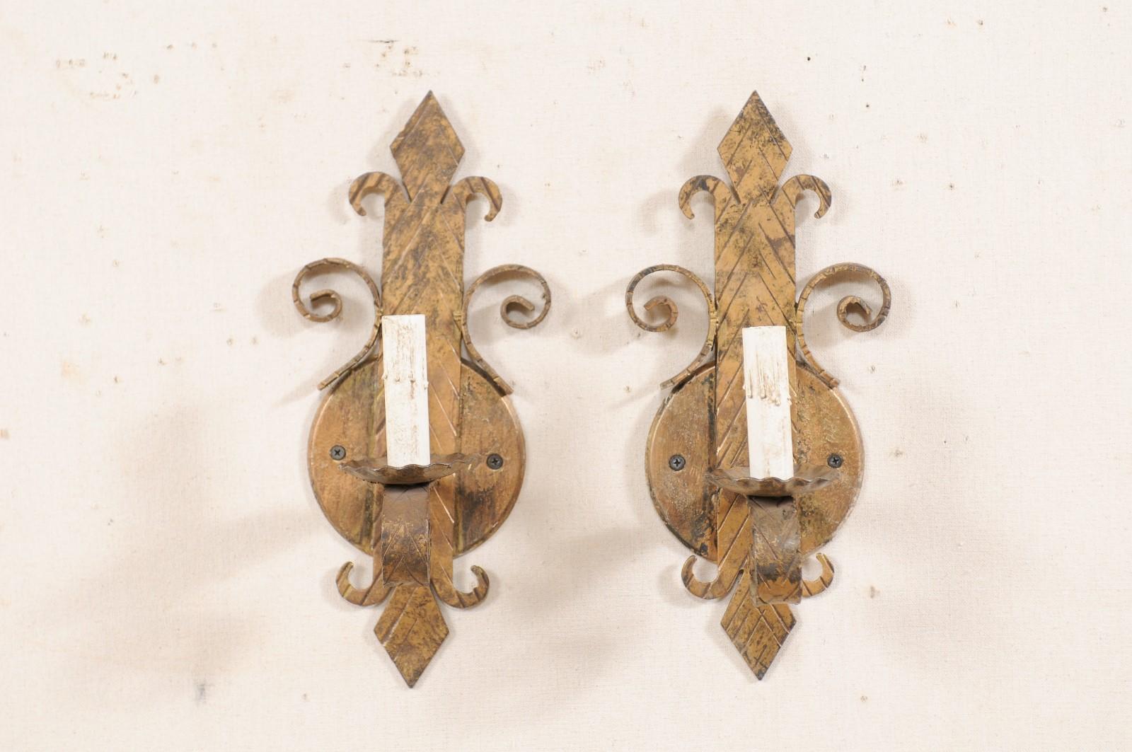 A pair of Spanish gold-tone iron sconces from the mid-20th century. This vintage pair of sconces from Spain each feature a stylized fleur-de-lys body with a single arm extending out into a downward curl, supporting a ruffled iron bobèche and painted