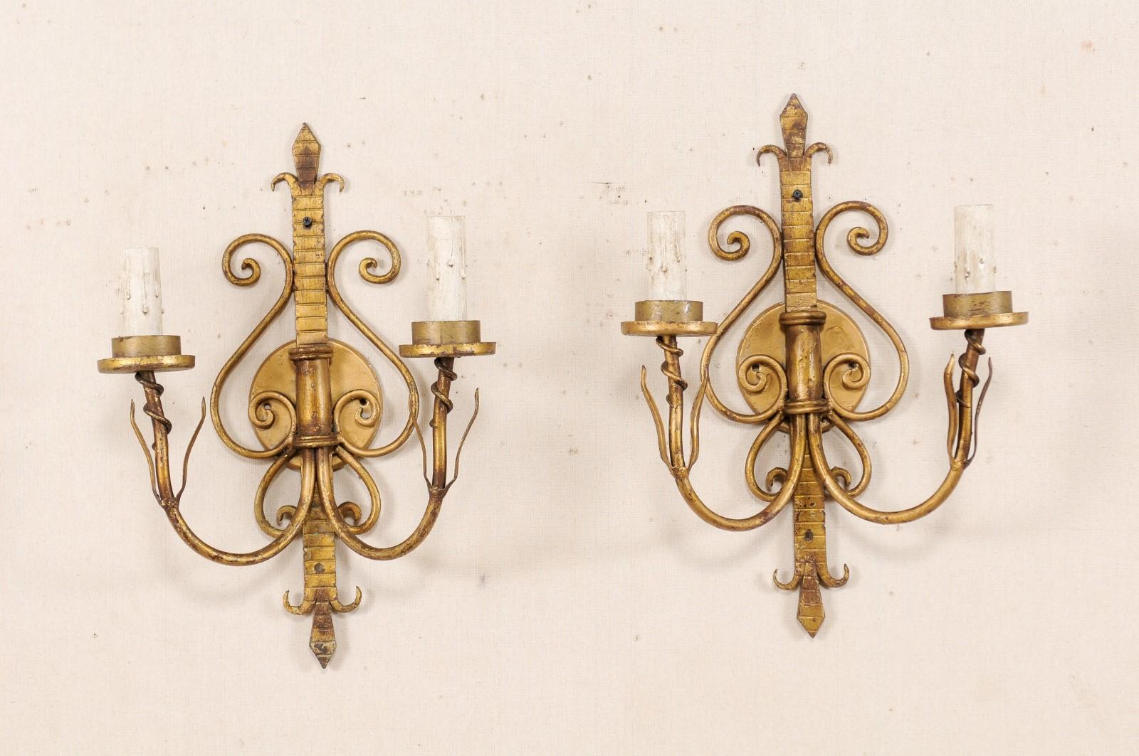 A pair of French two-light gold tone iron sconces from the mid-20th century. This vintage pair of French sconces each feature a vertically positioned and Fleur de Lys stylized and rib textured back above a rounded back-plate, with scrolling iron