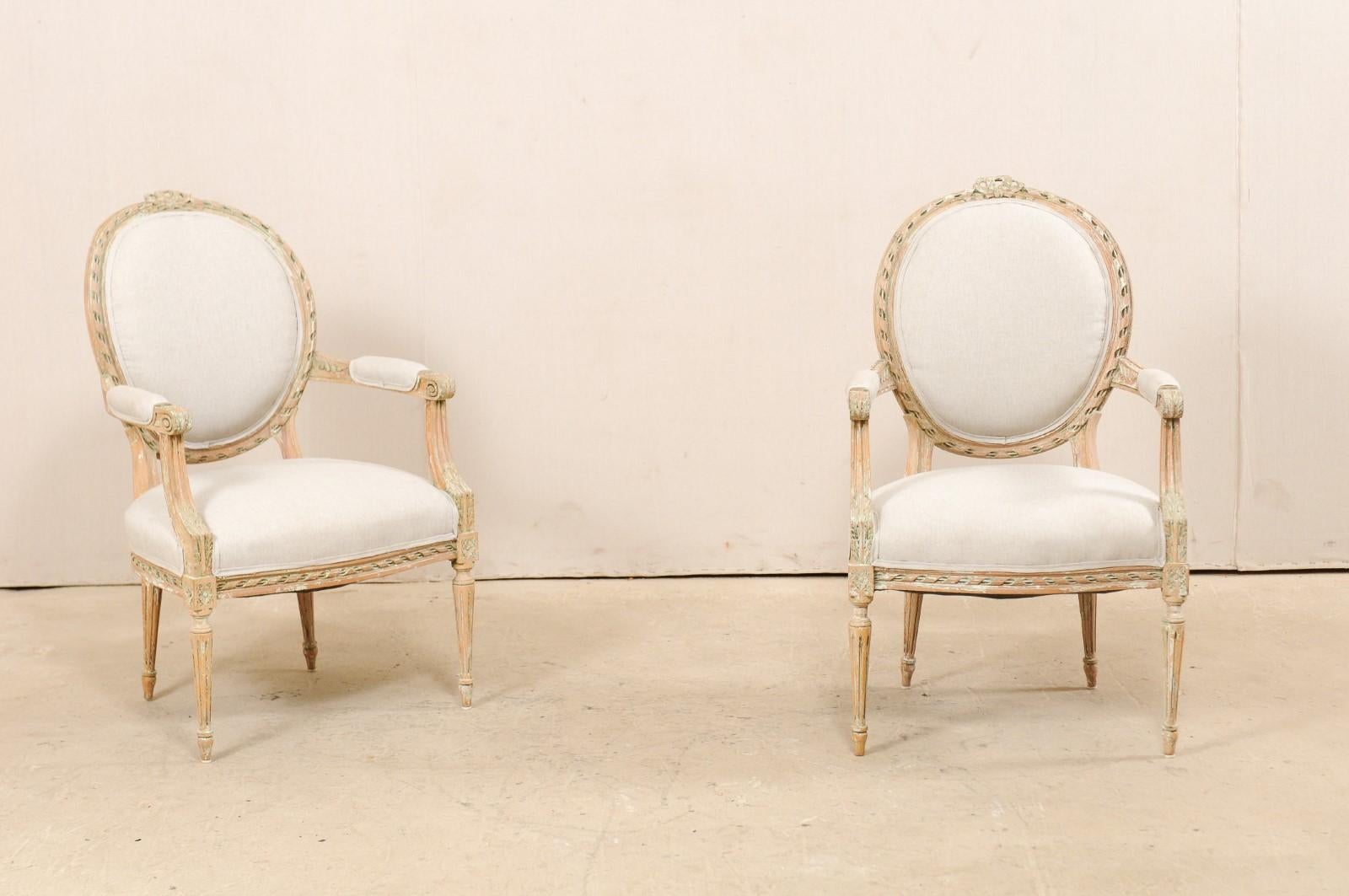 A pair of French carved wood and upholstered bergères chairs from the mid-20th century. This vintage pair of armchairs from France each feature oval-shaped upholstered backs with a wood frame accentuated with spiraled ribbon carved trim and pierced