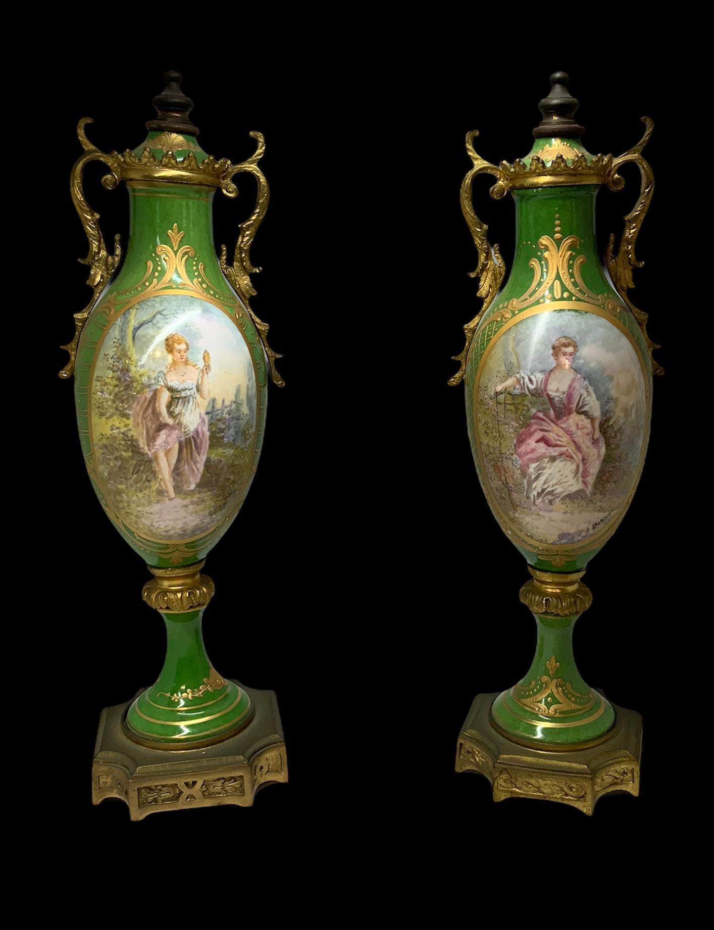 These are a pair of ormolu and green hand-painted and signed lidded urn vases. Each one depicting a scene of young ladies. One is sitting in a shore of a lake and looking at herself with a hand mirror and the other one is sitting on a stone bench in