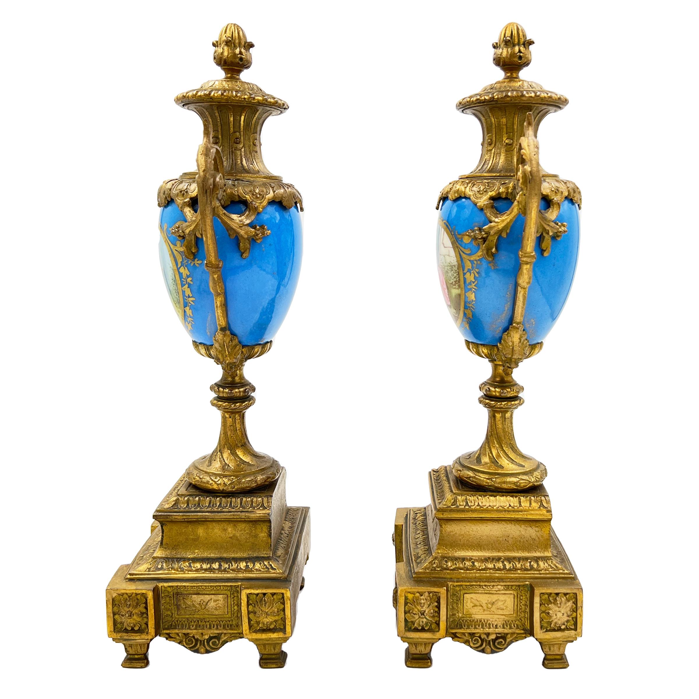 Exquisite pair of French Sevres-style porcelain urns adorned with gilt bronze mounts, featuring a celestial blue porcelain and idyllic rural scenes.
