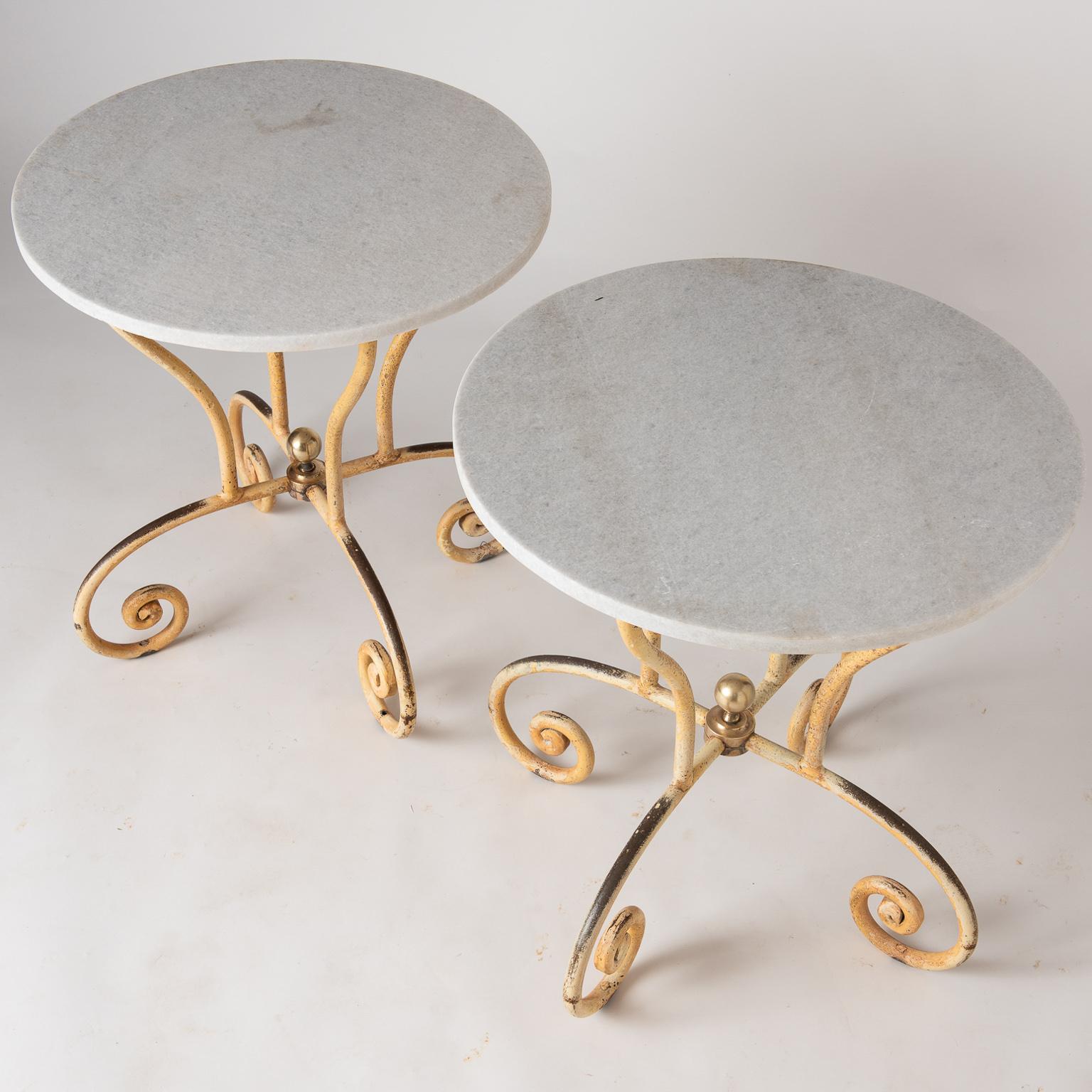 These chic, 20th century tables with curving iron bases in old yellow paint, are joined by a central brass fitting, topped with a brass finial. The round shaped marble is in perfect condition and the tables will work beautifully as cocktail