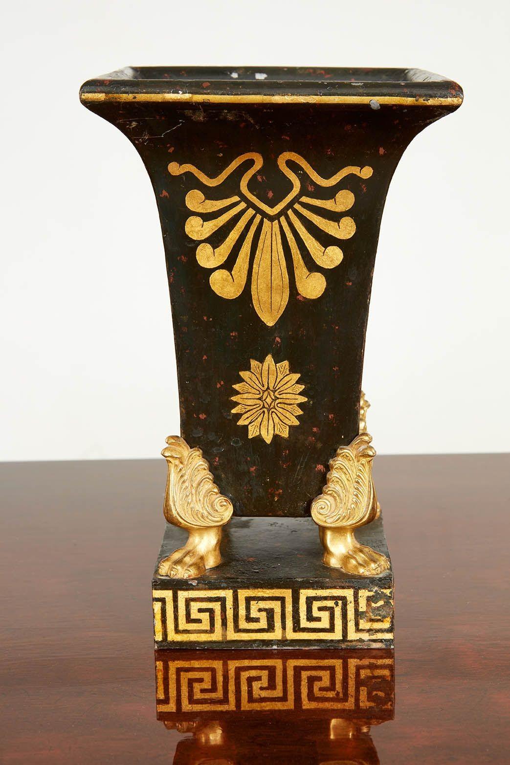 Coppia di vasi in toleware del periodo Impero francese con decorazioni nere e dorate, il corpo è decorato con anthemion e patera e poggia su piedi a zampa di leone con foglie d'acanto e su basamenti decorati con chiavi greche.