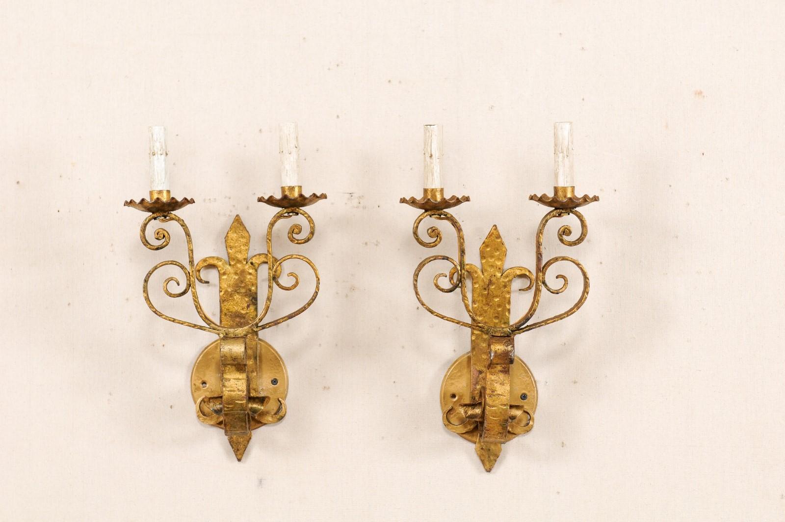 A pair of French two-light gold tone iron sconces from the mid-20th century. This vintage pair of French sconces each feature a vertically positioned and fleur-de-lys stylized back above a rounded back-plate. A thickly s-scrolled bar projects from