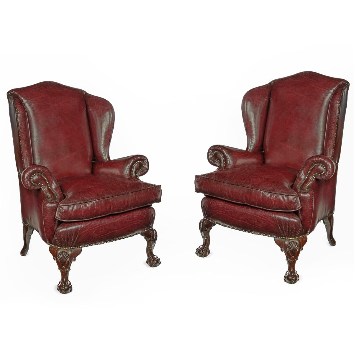 A pair of generous mahogany wing armchairs with shell carved knees, each with close buttoned scroll arms and generous seats, the legs carved with shells on the knees and ball and claw feet, reupholstered in distressed burgundy leather.  English,