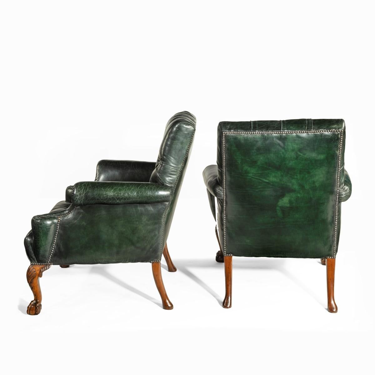 A pair of George II style walnut wing arm chairs, each with a generous back and seat with scrolling arms, reupholstered overall in deep green buttoned and distressed leather, the cabriole front legs carved with shells on the knees, the back legs