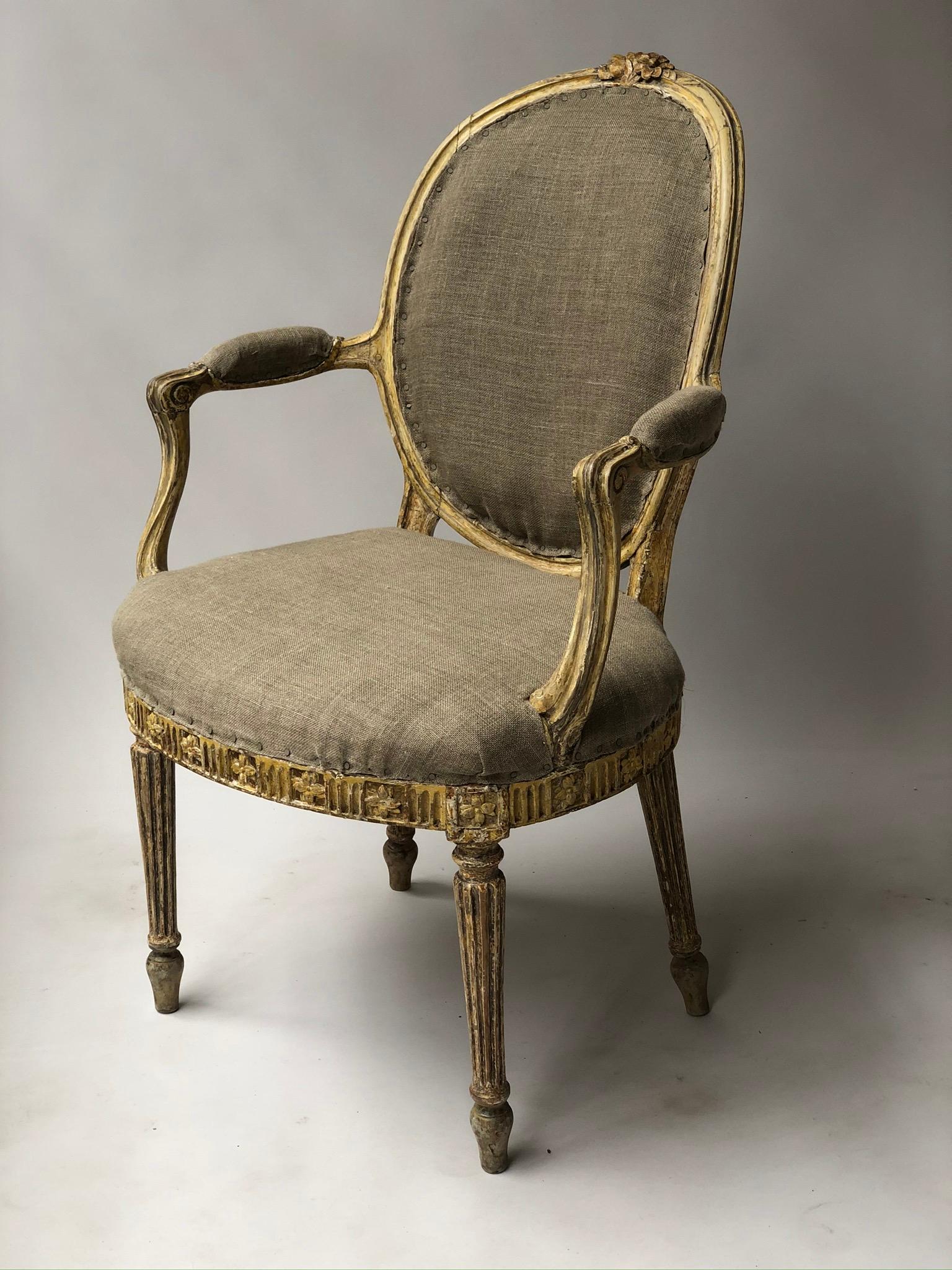 A pair of English George III period painted and gilded armchairs each with padded oval back, stuff over upholstered seat, carved and gilded frieze and turned fluted tapering supports
These circa 1770s English armchairs display much evidence of