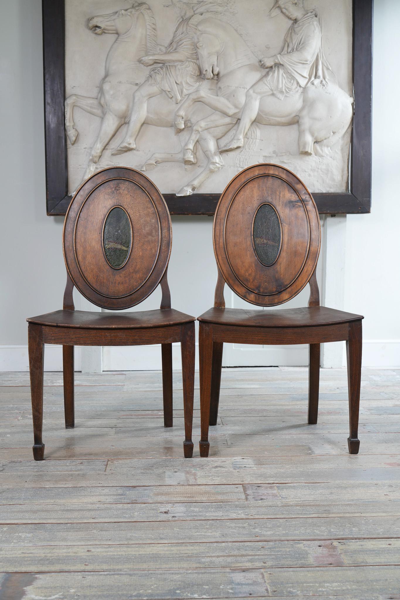  A pair of ash and elm side chairs in the manner of Mayhew & Ince.

The oval back centred by a crest, details below, above a shaped seat and square tapering legs with foot blocks, swept rear legs. The crest as depicted upon this Pair of George III