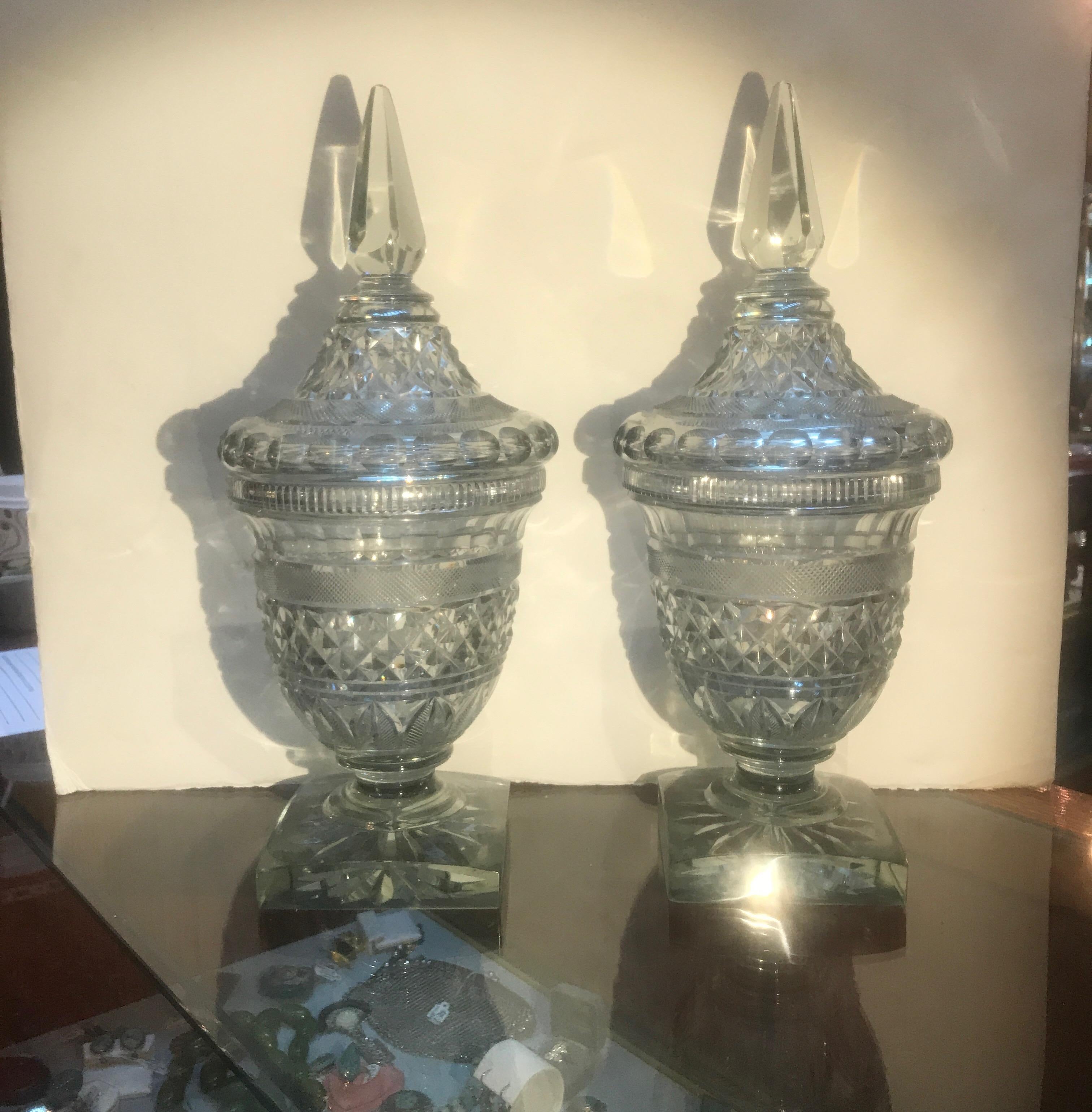 A pair of Georgian cut glass covered urns with spear finial tops. The faceted finials with domed cut lids with diamond cut bodies on pedestal bases with square bottoms.
