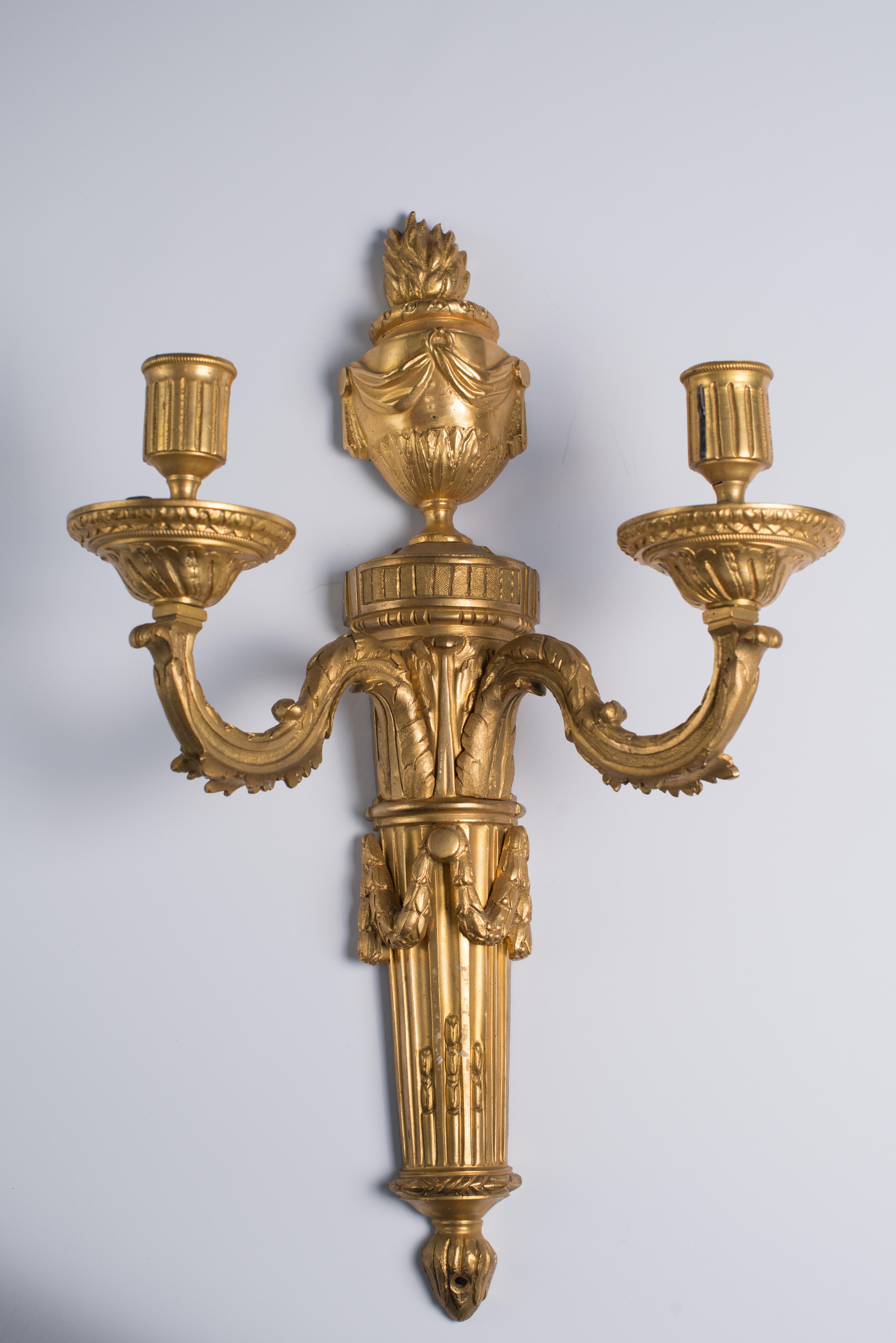 A pair of gilt bronze Louis XVI two-light wall sconces, France, circa 1780

Description
A pair of gilt bronze Louis XVI two-light wall sconces, each with a stop-fluted back-plate surmounted by an urn finial with drapery. Each sconce with two