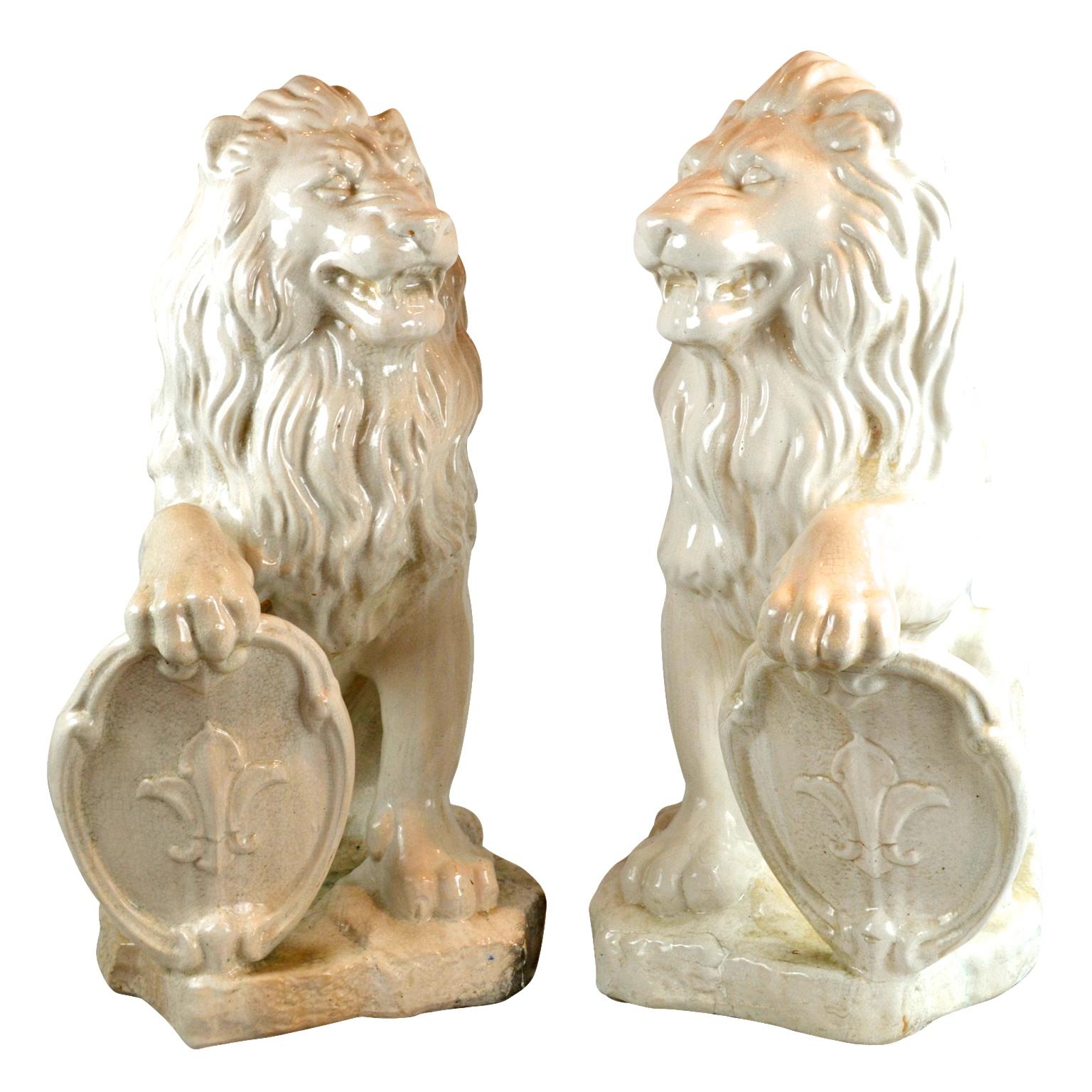 A decorative pair of terracotta rampant lions with white glazing, each sitting facing each other with one front foot resting on an oval shield decorated with a fleur-de-lys.

 