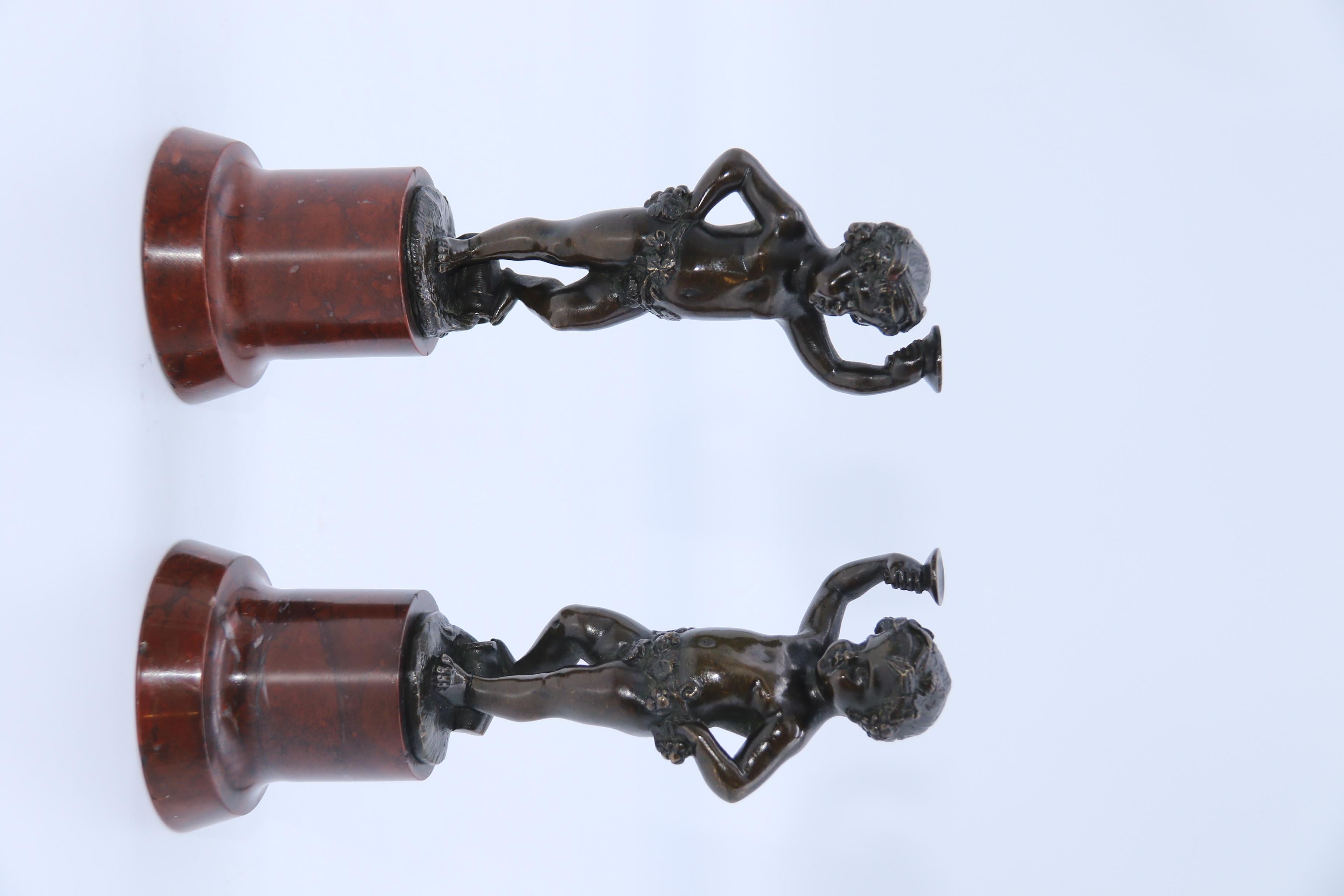 This small pair of fine Italian Grand Tour bronze figures are of classical putti naked apart from a belt of vine leaves and grapes around their lower torso. Each with one arm raised and holding a chalice of wine taken from the ewer which is lying on