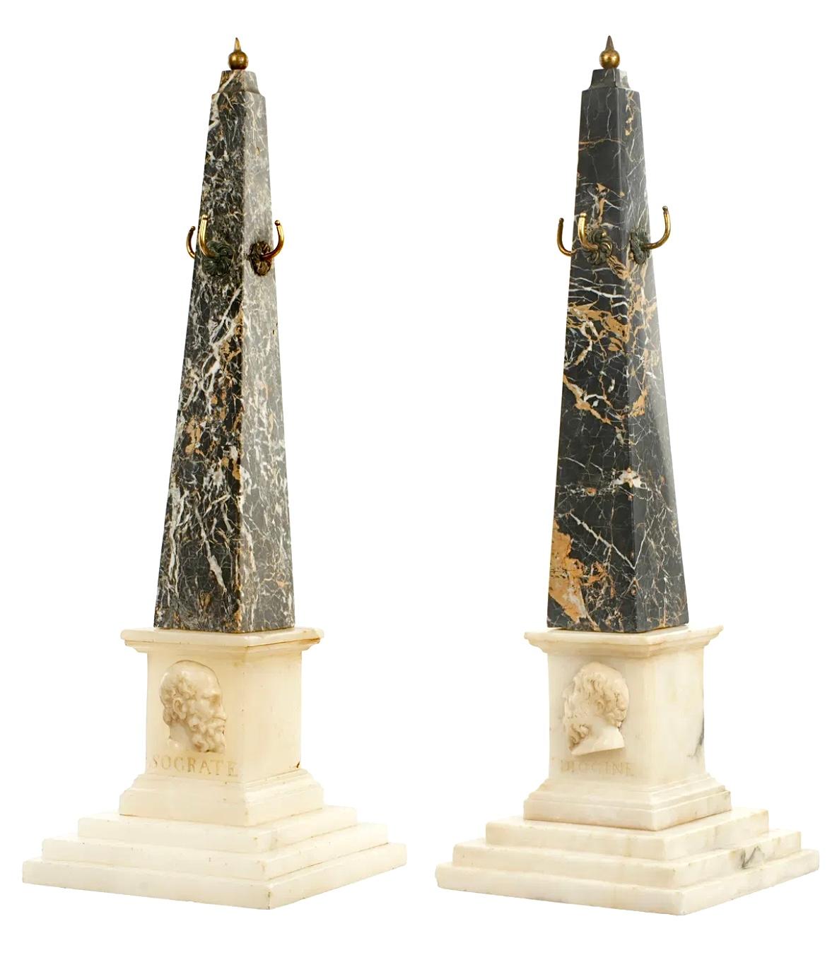 Coppia di Obelischi Grand Tour in marmo nero variegato del XIX secolo con terminali e ganci in bronzo applicati. Ognuno di essi presenta i profili in rilievo dei filosofi greci. Una con busto di Diogene su base di marmo a gradini, l'altra con busto