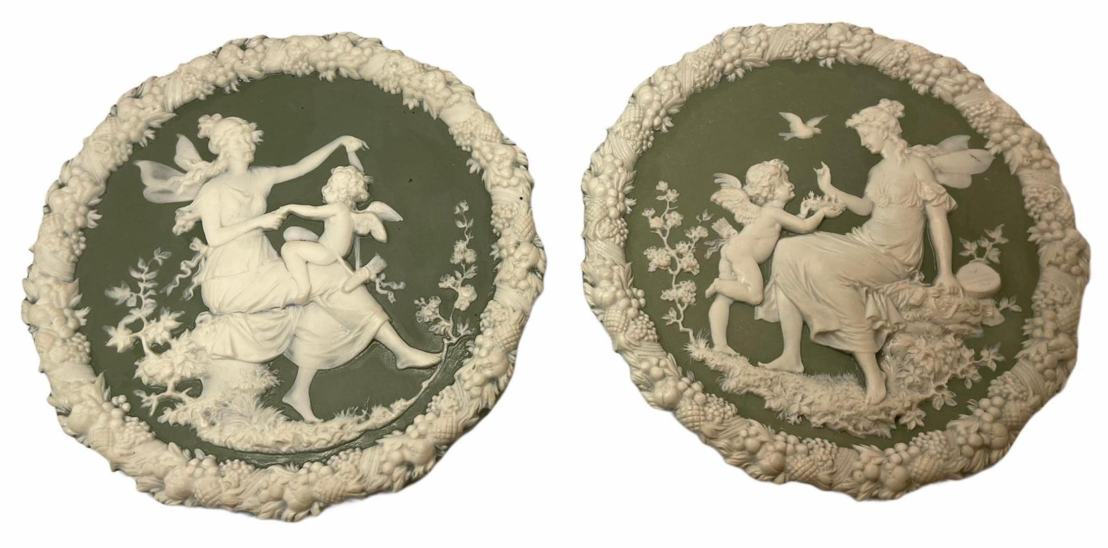 These are two green and white jasperware plaques. One is depicting a decorative relieve scene of a nymph holding hands with a cherub in her lap and playing with him. In the other one the nymph is holding a banjo and observing a cherub that is