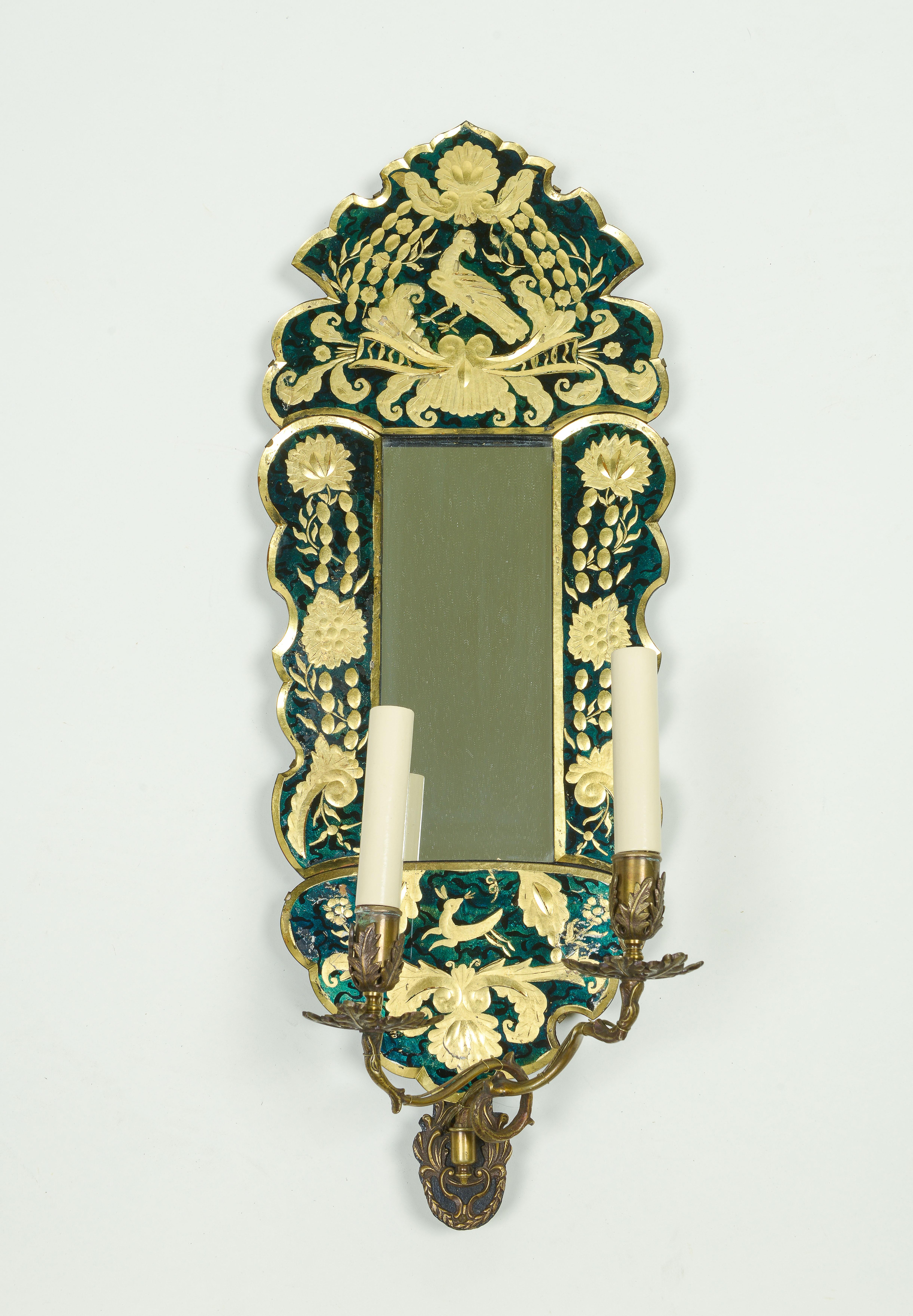 Each with rectangular mirror plate within a green ground verre eglomise scalloped and arched surround overpainted with gilt decoration incorporating birds, flowers and foliage; issuing two brass electrified candlearms with ivory candle sleeves.