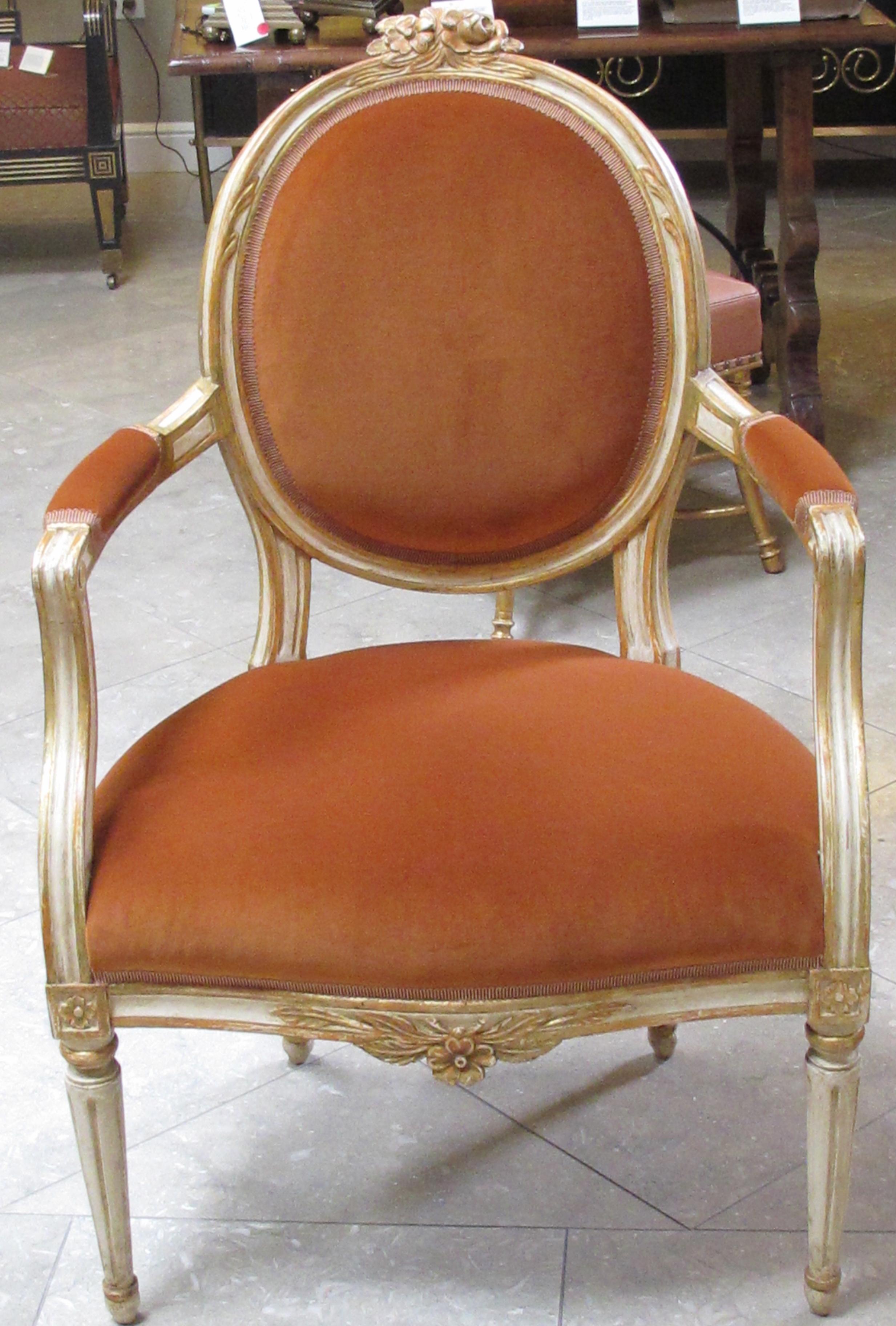 An elegant pair of Swedish Gustavian style ivory painted and parcel-gilt oval-back armchairs; each arching crest surmounted by a floral crest above an oval padded back; joining a serpentine tight seat flanked by padded arms; raised on fluted turned