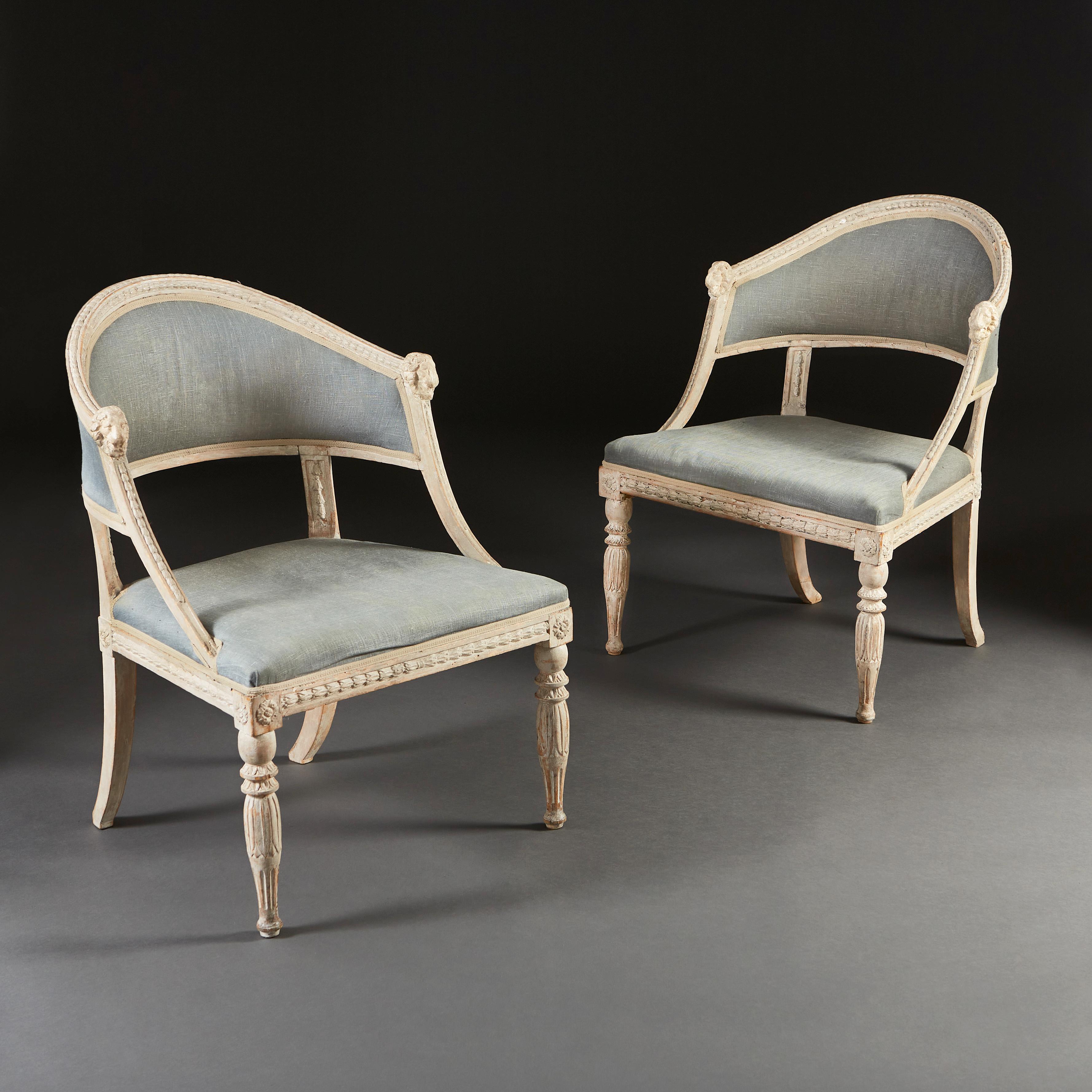 A pair of early nineteenth century Gustavian Swedish armchairs with carved and painted frames, with curved backs carved with laurel leaves and terminating with masks to the arms, upholstered in blue linen.