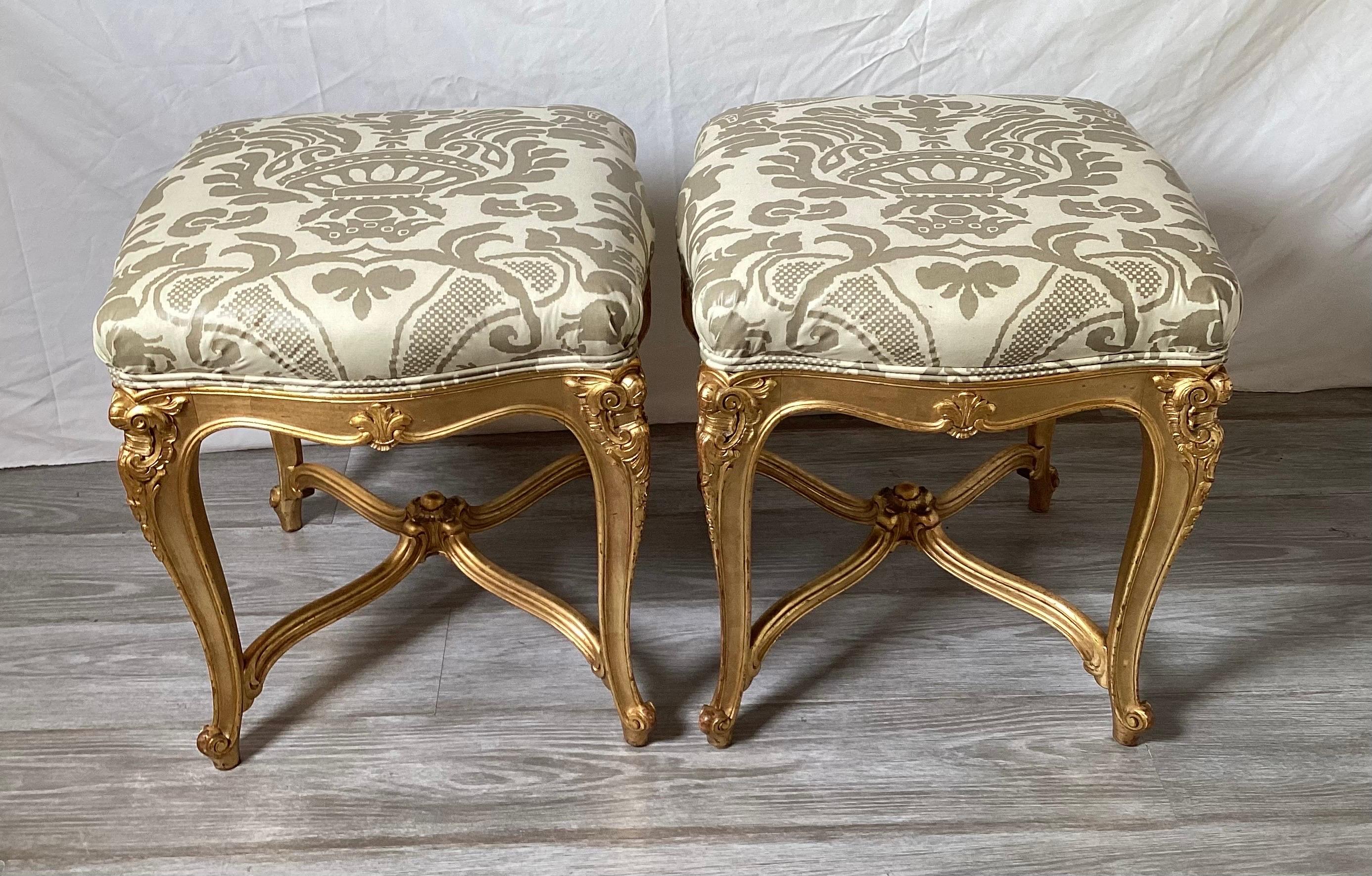 An Elegant pair of hand carved gilt wood square benches with new fabric. The real gold leaf frames with hand carved details with an X stretcher base. The new upholstery is vintage Fornisetti style champagne and taupe damask print.