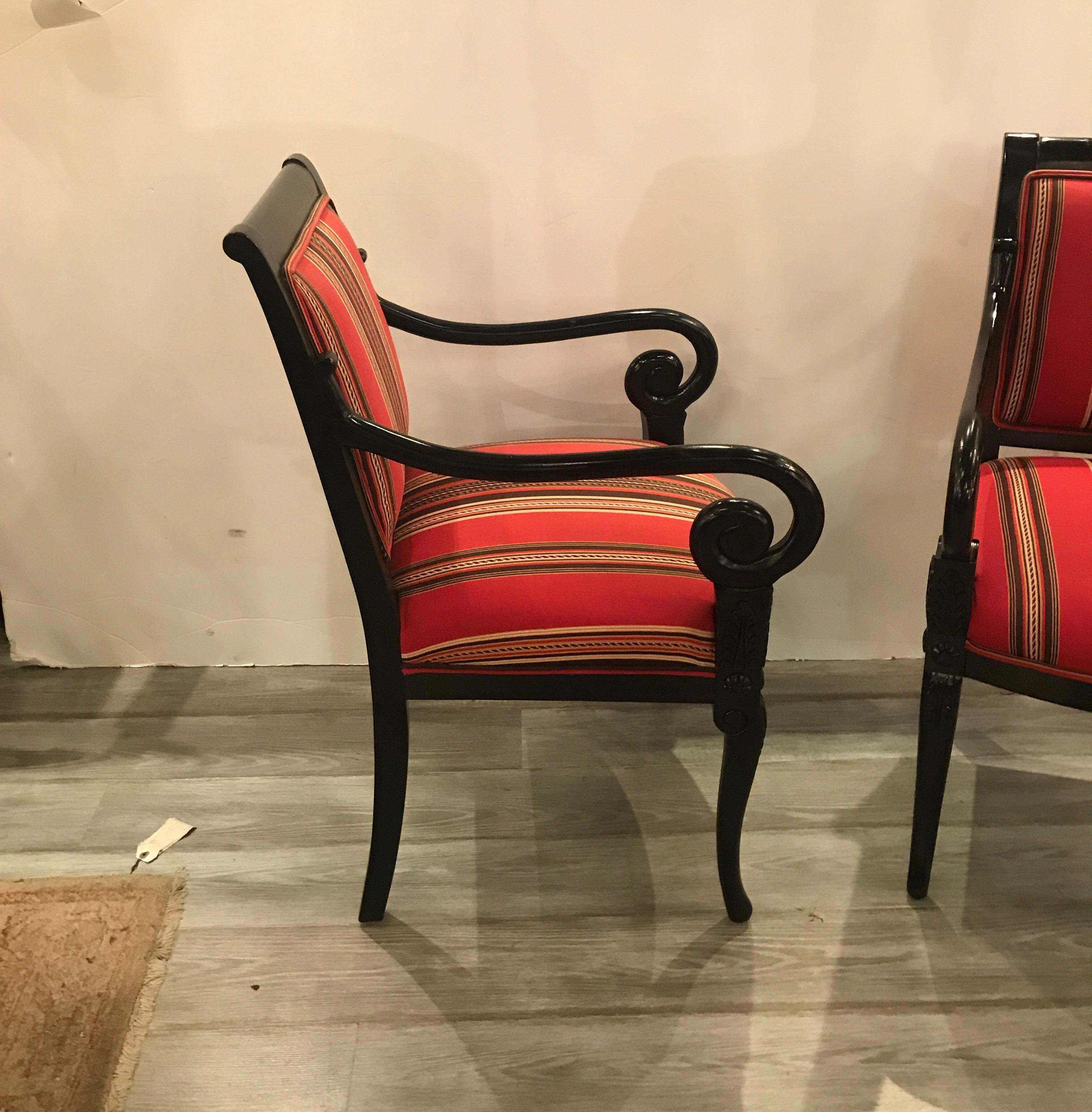 A stylish pair of Hollywood Regency neoclassical armchairs with new upholstery. The black ebonized frames with carved detail on the upper part of the front legs with graceful scrolling arms. The back and seats in a vibrant woven red fabric.