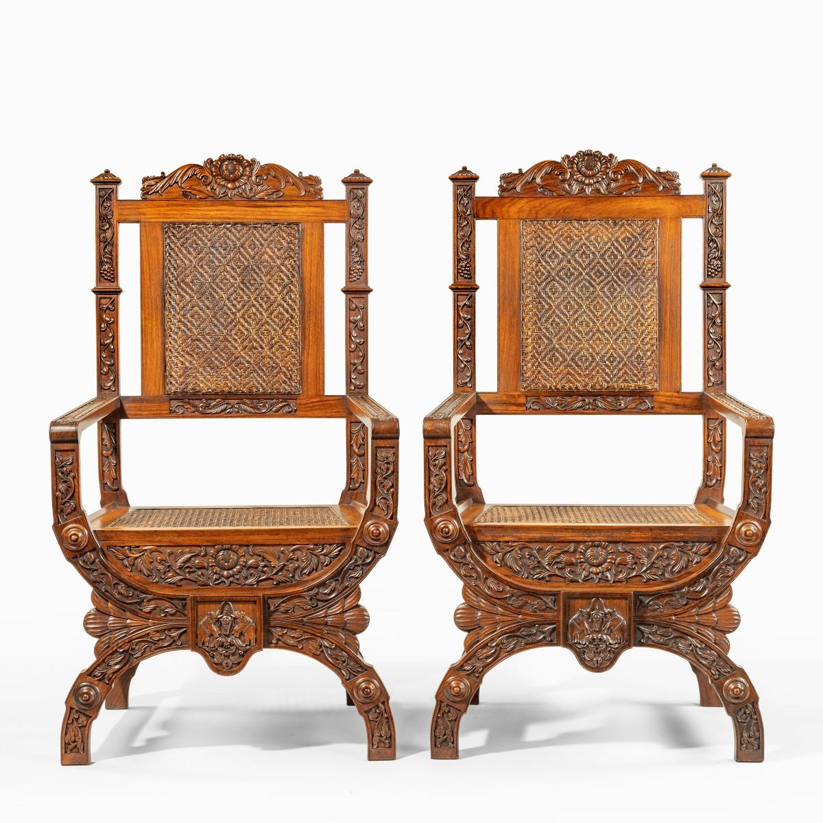 A pair of Indian throne chairs, carved with the arms of the Kingdom of Travancore, each of curule or “x-framed” form carved with fleshy naturalistic flowers, tendrils, vines and foliage, the seat and back with rattan panels, the X centred on an