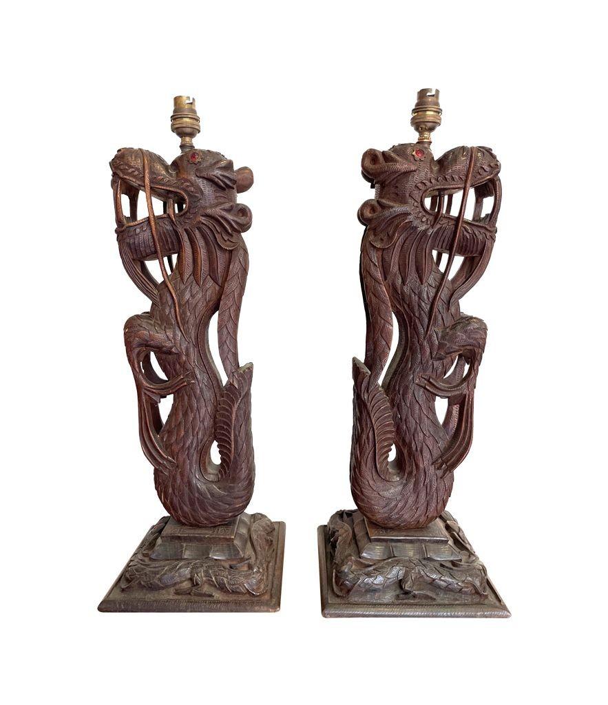 Ein Paar kunstvoll geschnitzte burmesische Holzlampen mit jeweils einem aufrecht stehenden Drachen auf einem geschnitzten Holzsockel, um den sich ein weiterer Drache schlängelt. Beide Drachen haben rote Steinaugen. Neu verkabelt mit neuen
