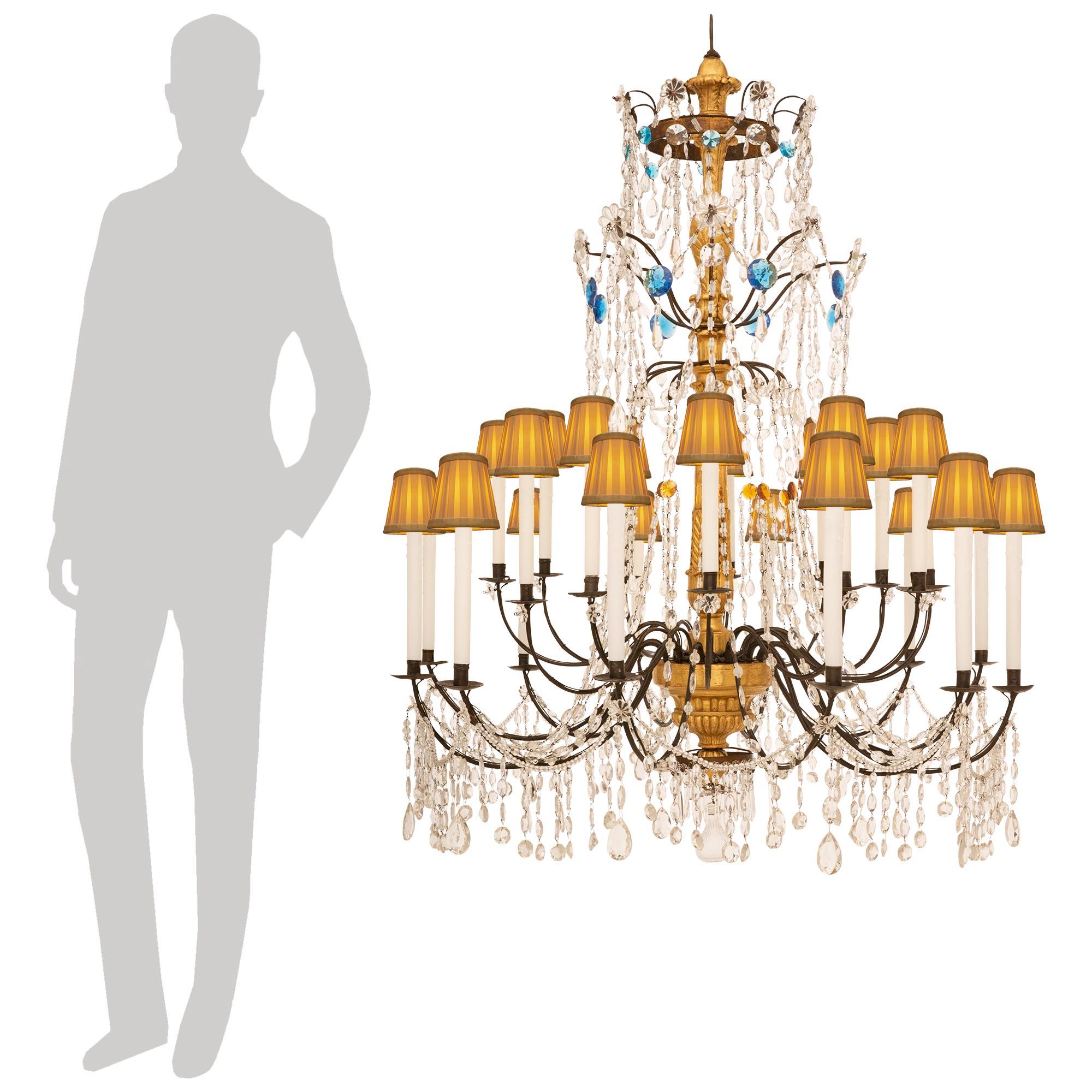A stunning and most elegant pair Italian 18th century Genovese st. Giltwood, colored Glass and Crystal chandeliers. Each most decorative twenty four light chandelier is centered by a wonderful circular reserve surrounded by prism shaped Crystals and