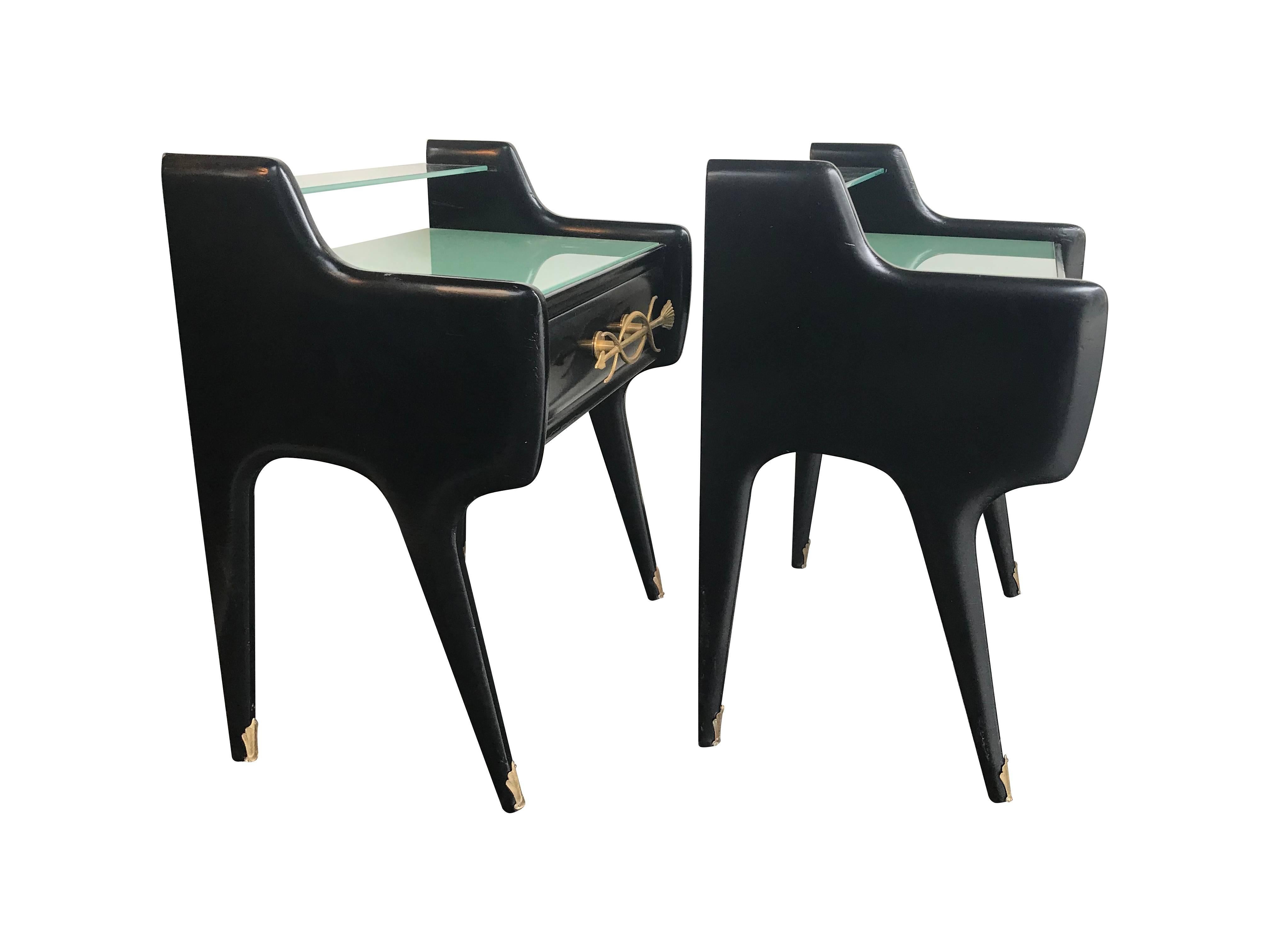 A pair of Italian 1950s ebonized bedside tables with a single drawer in each, with ornate arrow motif, gilt metal handles. Each with green frosted glass top, clear glass top shelf and brass ormolu leg plates.