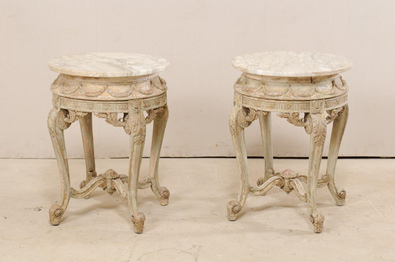 A pair of generously carved, Rococo inspired accent tables with marble tops from the mid-20th century. This vintage pair of American tables, created in Italian design, feature round marble tops which overhand their heavily carved, rounded bodies