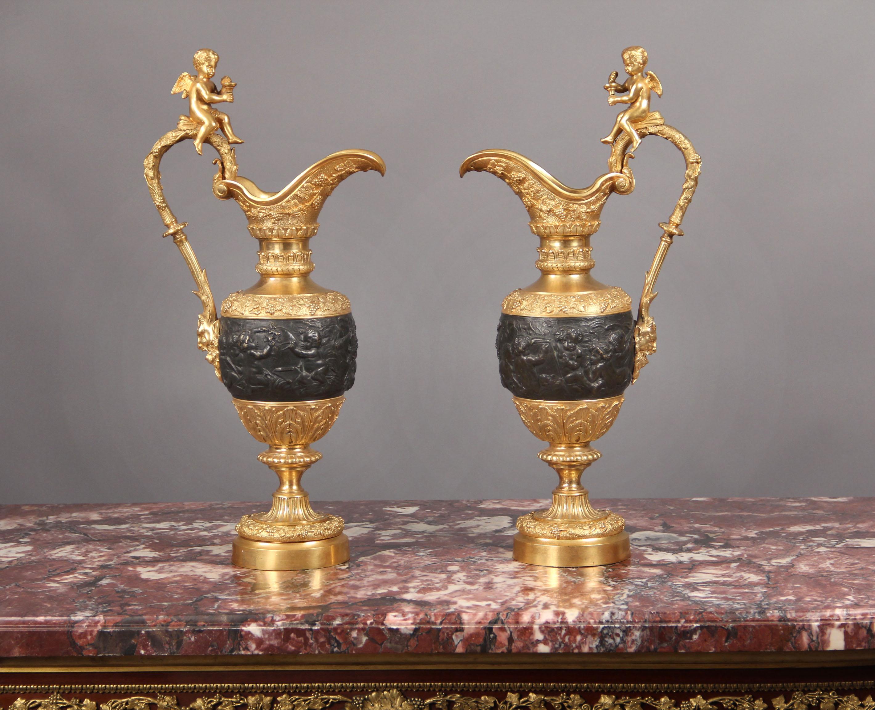 A pair of late 19th century gilt bronze and patinated bronze ewers.

The scrolled lip above a slender neck and bulbous body cast with Bacchic putti, the scrolled handle mounted with a seated putto and satyr mask.
