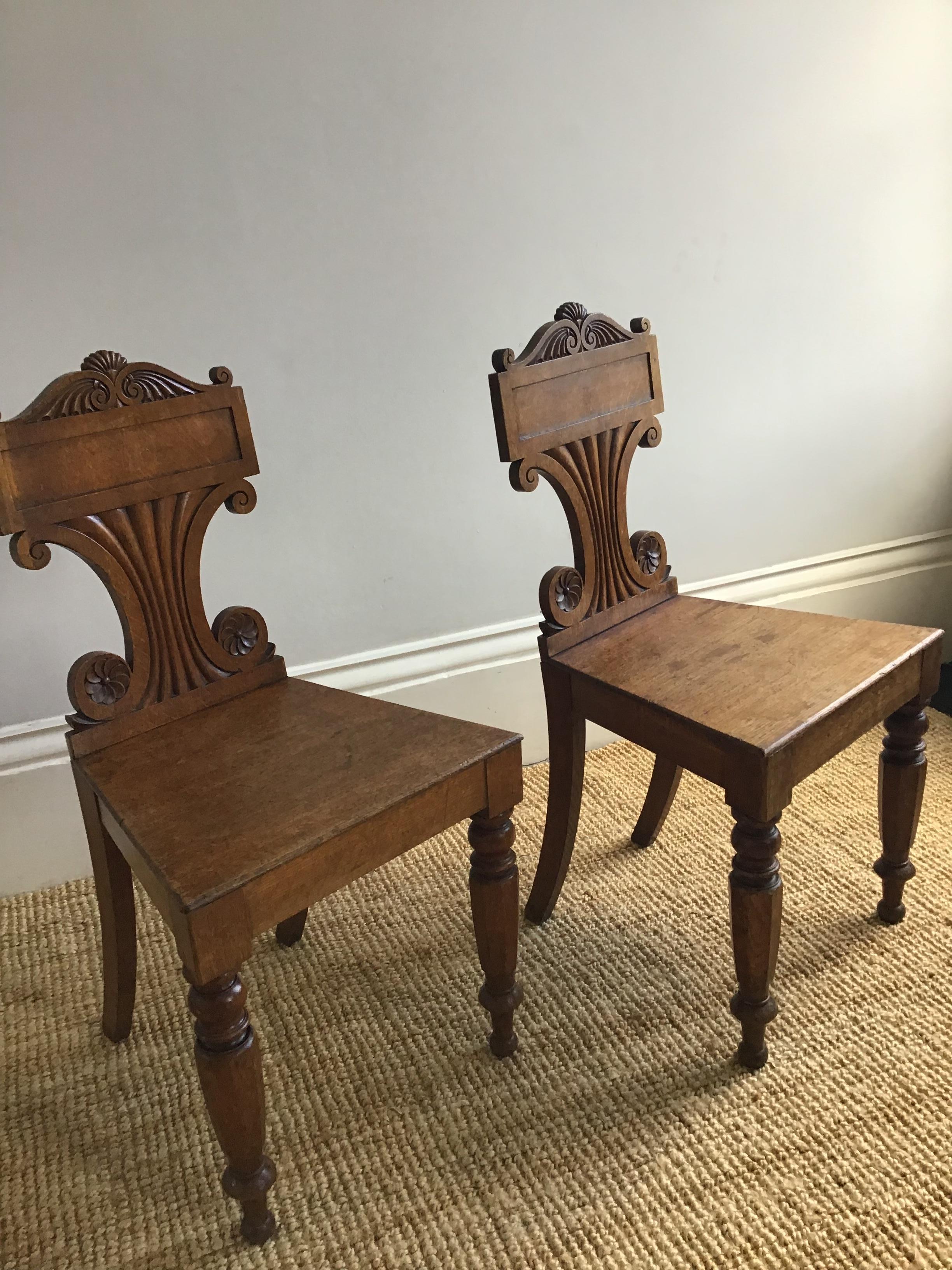 A pair of English late Regency period carved oak hall chairs with carved scroll crest, tablet centre, waisted back,
panel seat and faceted turned front supports
Reflecting at the time of their making a move away from imported mahogany to an