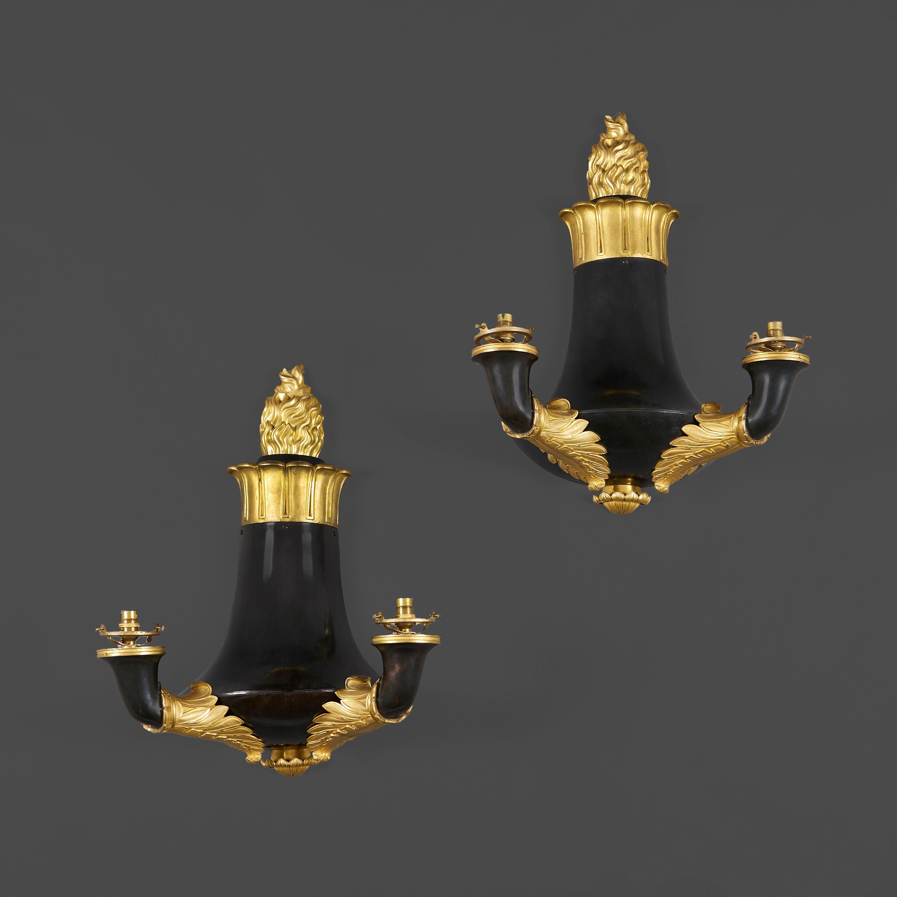 Pareja de apliques de bronce y ormolina de estilo Louis Philippe y diseño Colza, el cuerpo abocinado y patinado bajo una corona de ormolina de hojas estilizadas coronada por flambé, que sostiene dos ramas en forma de cuerno con cuellos de