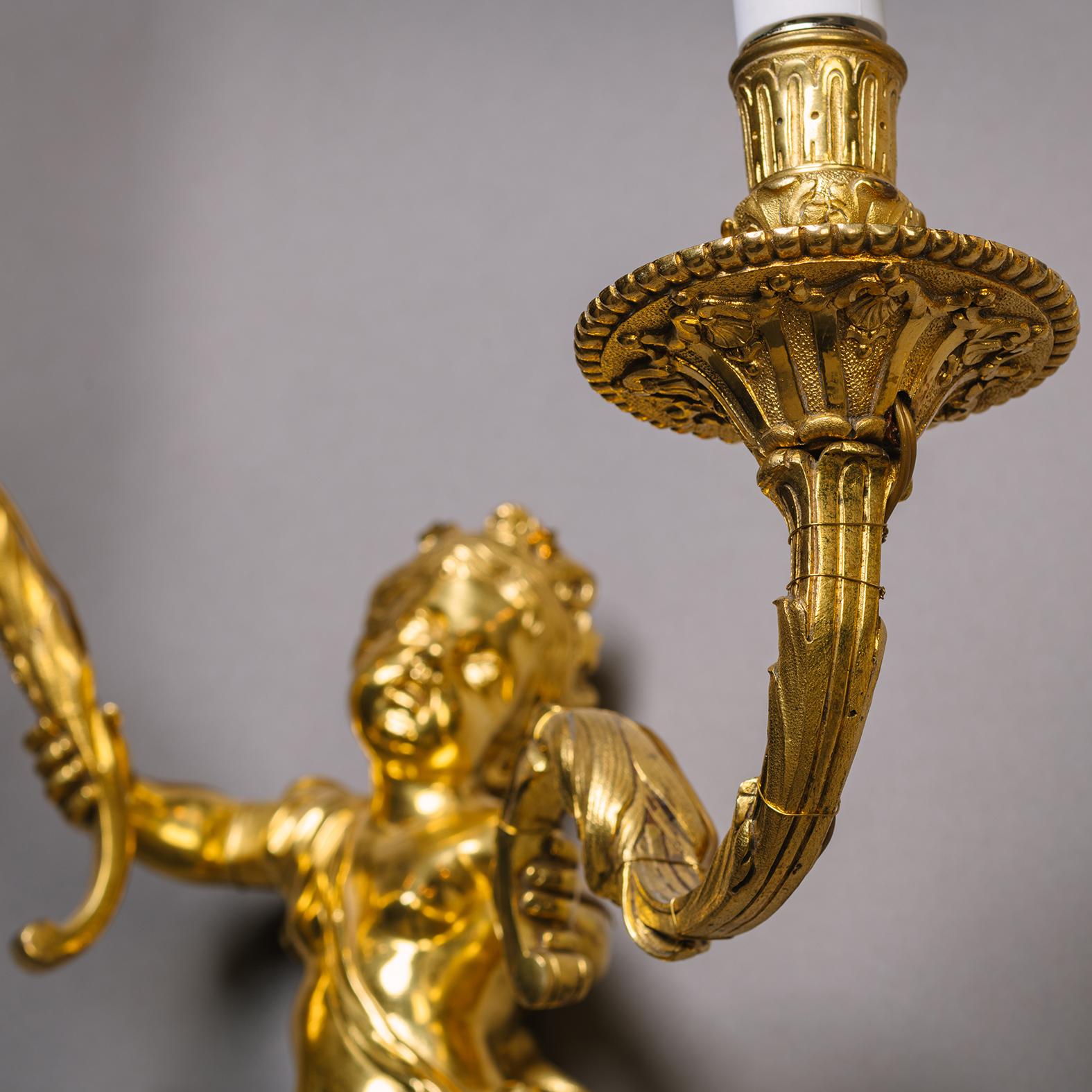 Ein Paar vergoldete Bronzewandapplikationen im Louis-XIV-Stil mit zwei Leuchten.

Jedes Modell stellt einen Cherub dar, der aus einem mit Blattwerk geschmückten Bügel herausragt und einen 