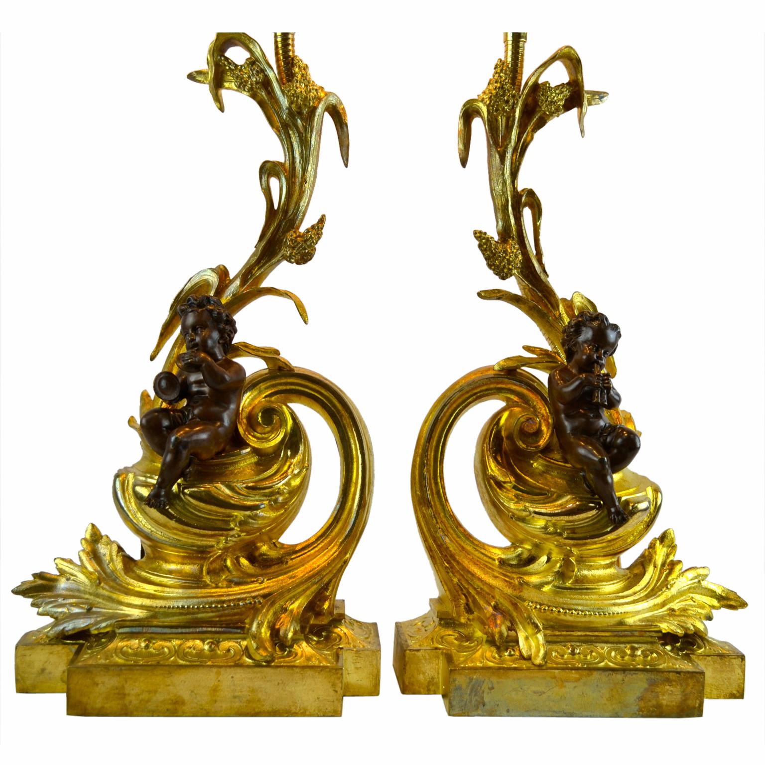 A pair of lamps made out of Louis XV style gilt and patinated bronze of chenets or fireplace andirons) The elaborate gilt bronze Rococo scrolling bases are entwined by seated patinated bronze cupids each playing a musical instrument. The upper