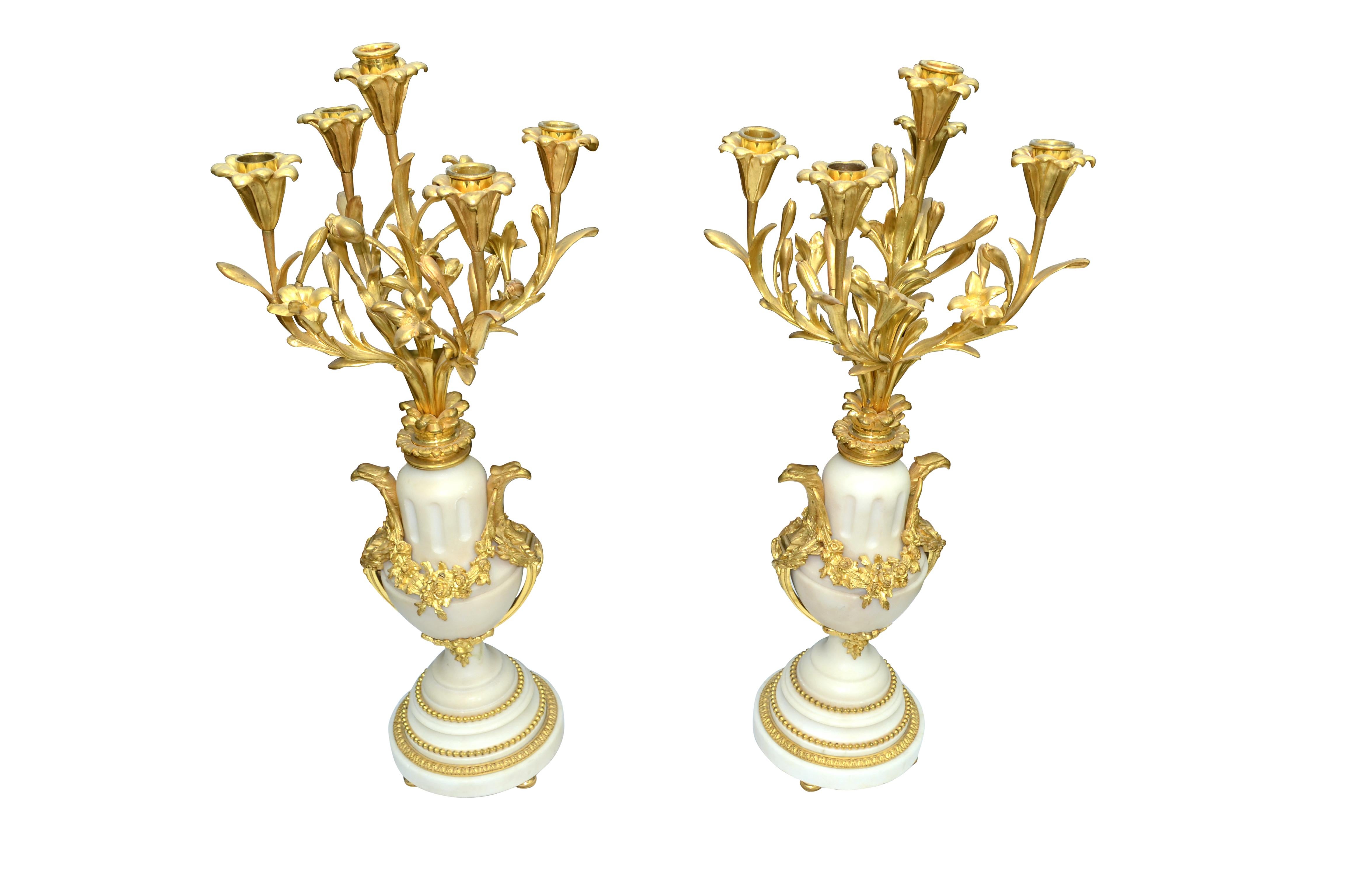 The white marble bodies of the candelabra are urn shaped and sit on circular stepped marble bases on gilt ball feet. On either side of the urns are stylized eagle head 'handles' which are joined along the wasted body of the urn with finely chased