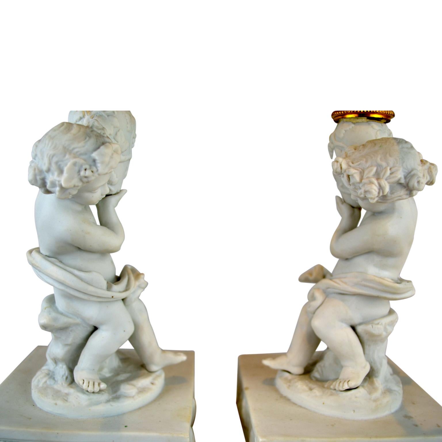 A pair of figural bisque porcelain (unglazed porcelain), candelabra each featuring a partially draped nude cupid sitting on a tree trunk holding aloft an urn which supports two curved candle arms; the whole on a rectangular bisque bases on a further