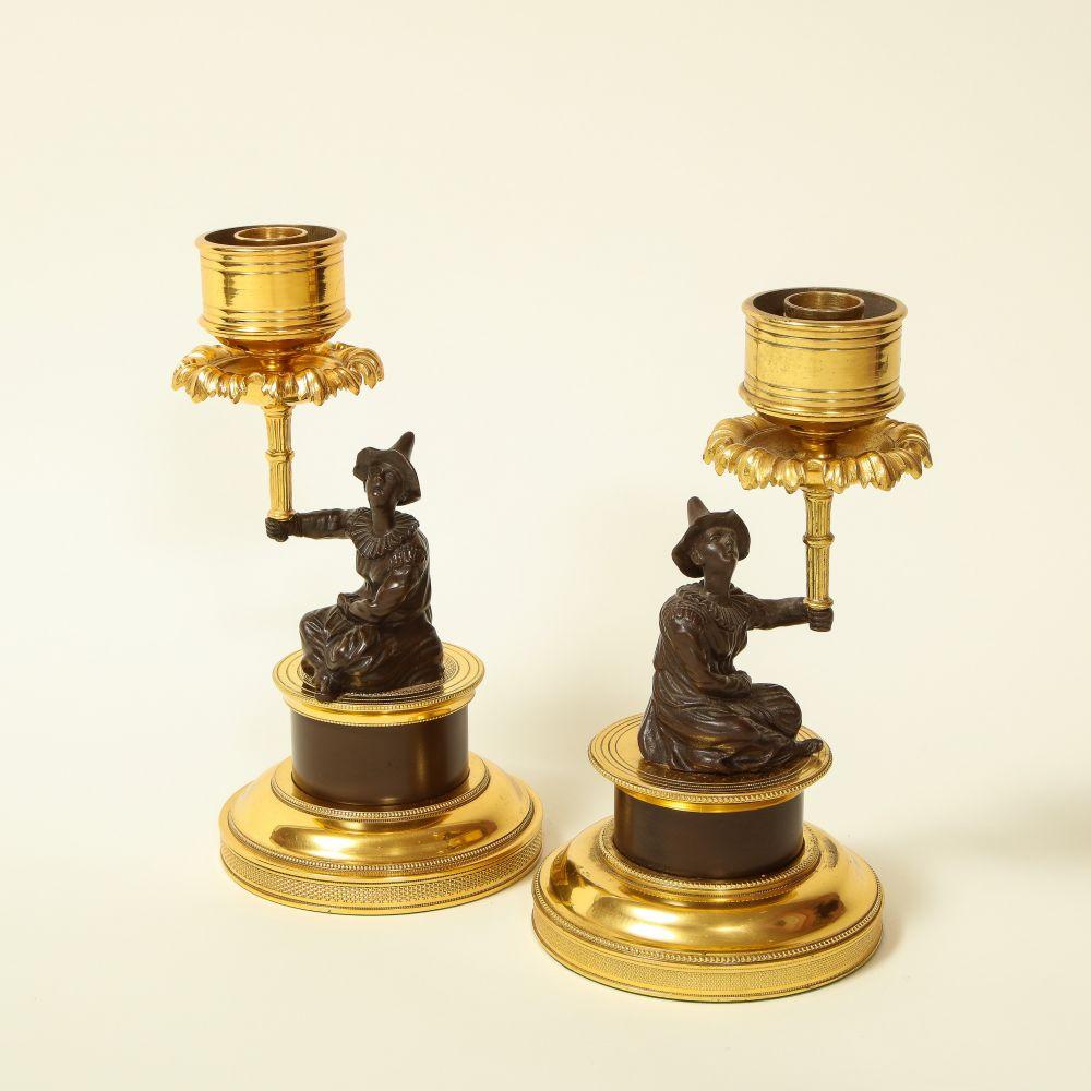 Each with a bronze figure of a seated harlequin dressed in the 18th century manner seated on a turned matte bronze base with gilt bronze plinth and with outstretched arm holding a stylized palm frond beneath the candle nozzle.

From the Collection