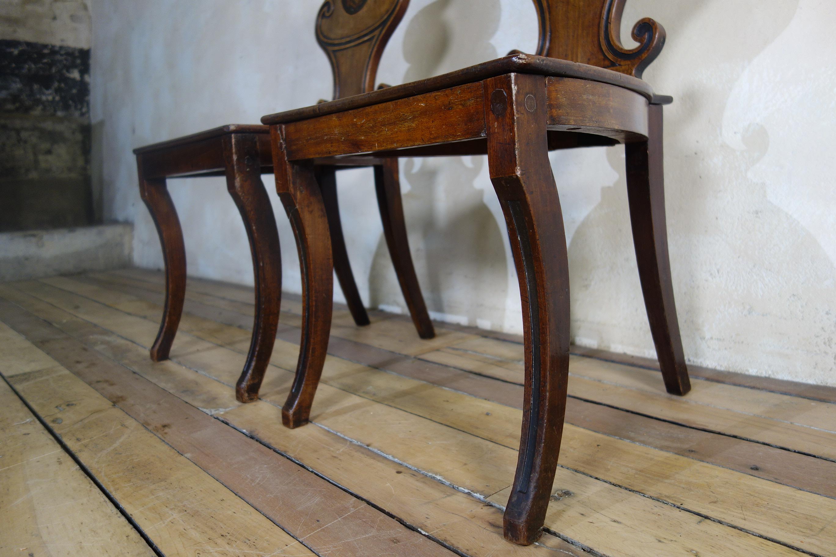An exceptional pair of George III mahogany hall chairs.  Demonstrating an Italian influence displayed in the single ebonized fluted decoration to the scrollwork design and front sabre legs. Presenting a rich and warm patination