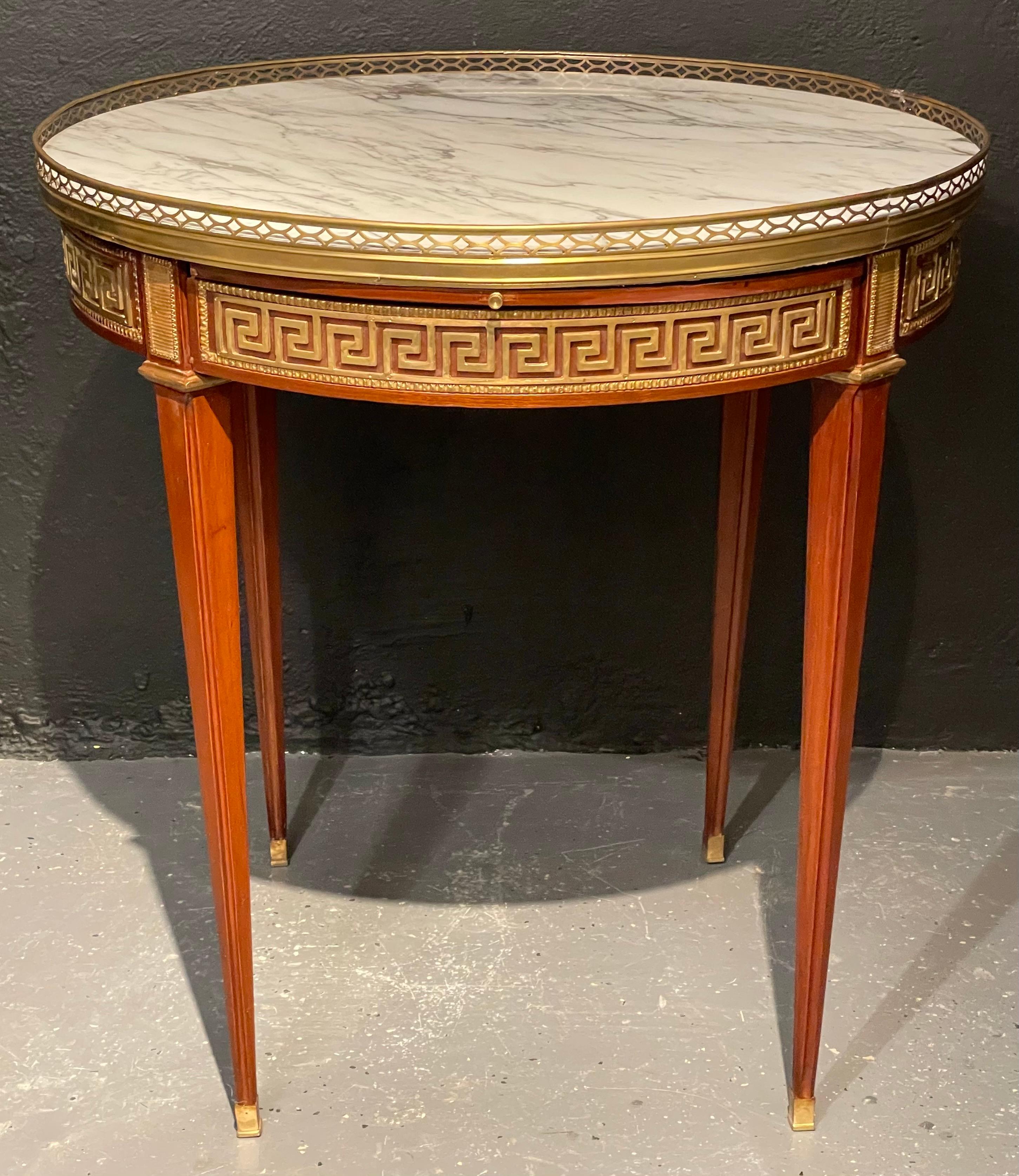 A pair of marble top Greek Key Bouillotte or end tables. Manner of Jansen in mahogany with double drawers and pull-out slides. Each bronze sabot leading to a group of tapering legs terminating in bronze capitals supporting an apron have full Greek