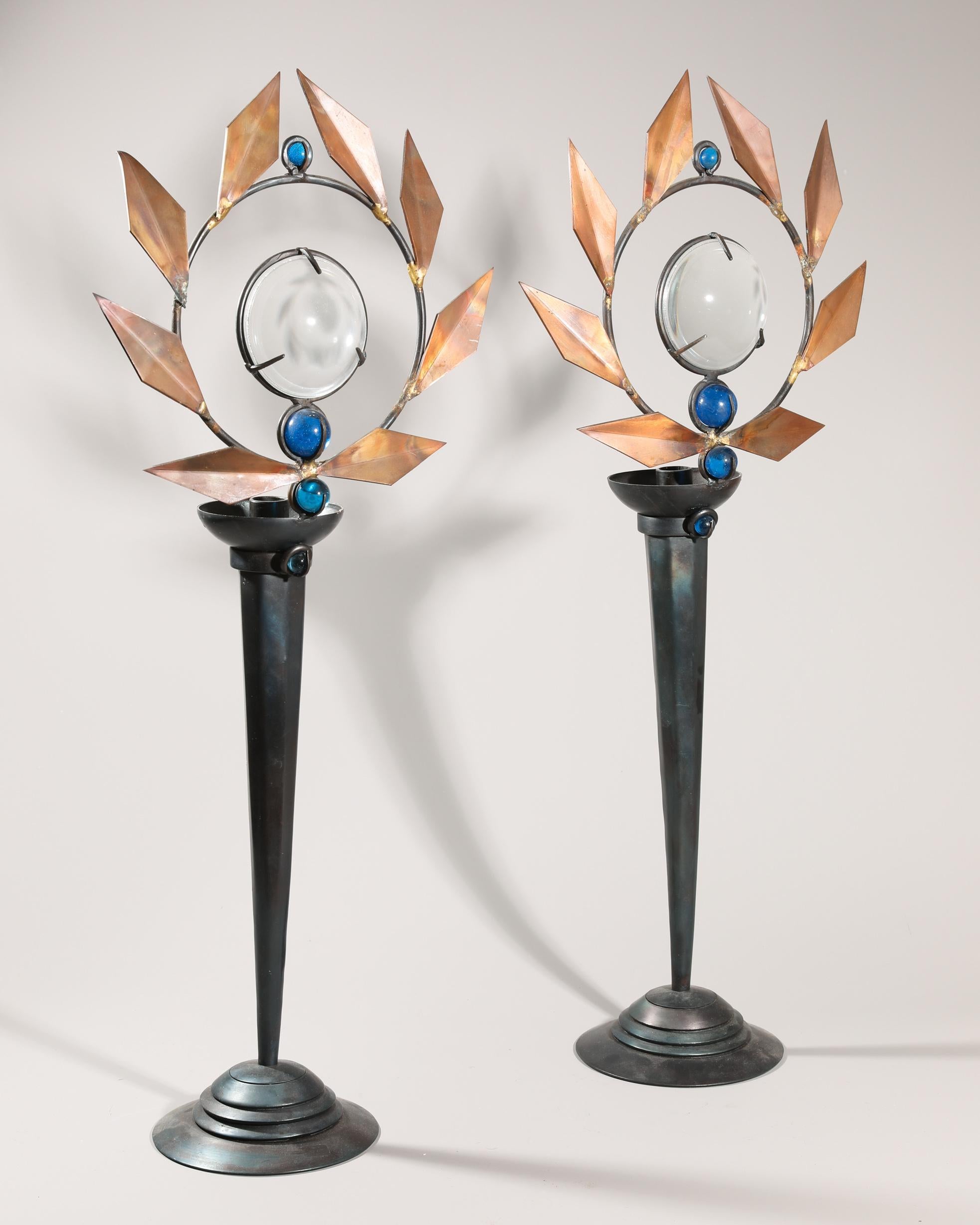 Una coppia di candelieri disegnati da Marc Brazier-Jones nel 1986 circa, modello Olympia. Realizzato in lamiera d'acciaio patinata e rame, il design presenta una base affusolata che si innalza verso una cornice circolare ornata da foglie angolari in