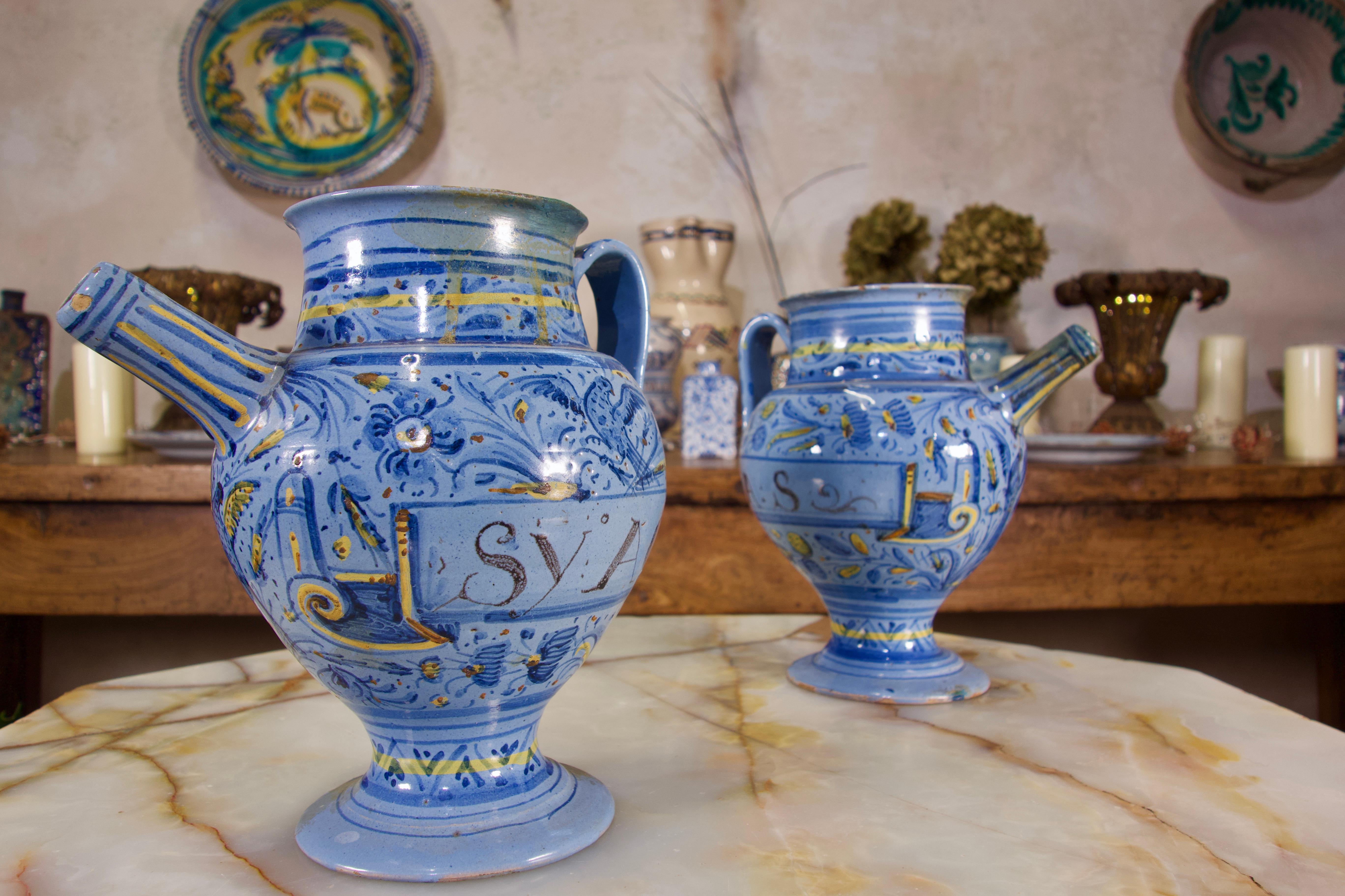 A pair of mid-17th century Italian maiolica Berettino syrup or wet drug jars. Demonstrating bulbous bodies inscribed 'MEL:ROSA:S' and 'SY:ACETOS.DIAROD' within painted labels against a ground of flowers and foliage picked out in yellow and ochre.