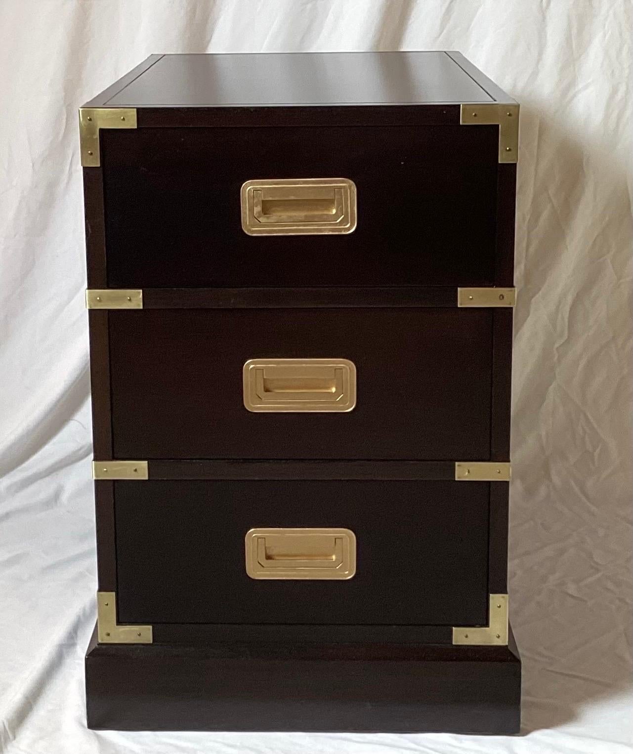 A Pair of chic dark walnut stained campaign style end tables or side chests by Baker Milling Road. The dark cabinets, with polished brass mounts and drawer pulls in a timeless campaign style. The chests with three working drawers with brass corners